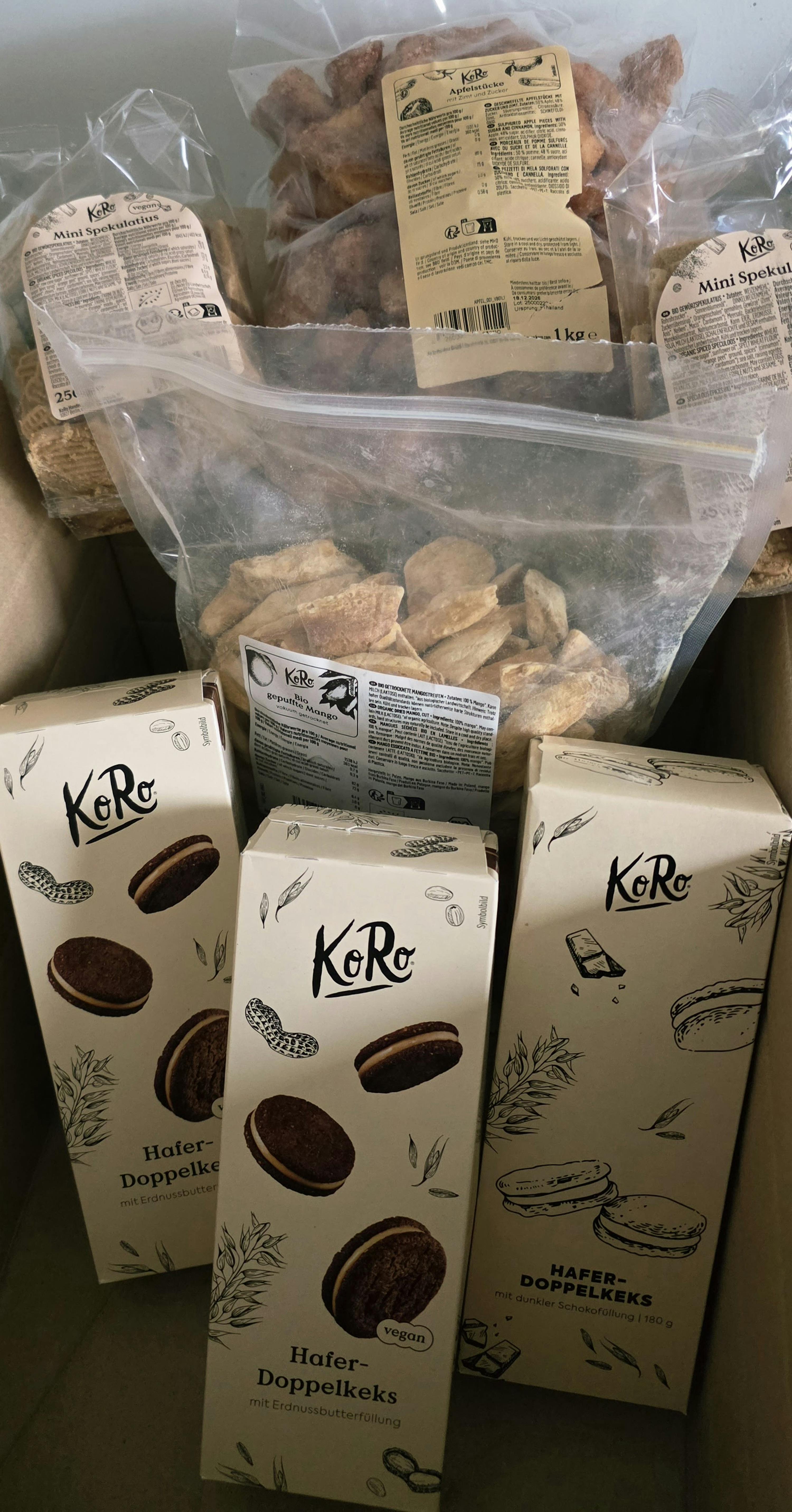 eine nahaufnahme einer kiste voller koro-produkte, darunter drei schachteln hafer-doppelkekse, tüten mit apfelstücken, mini-spekulatius und bio-mango.