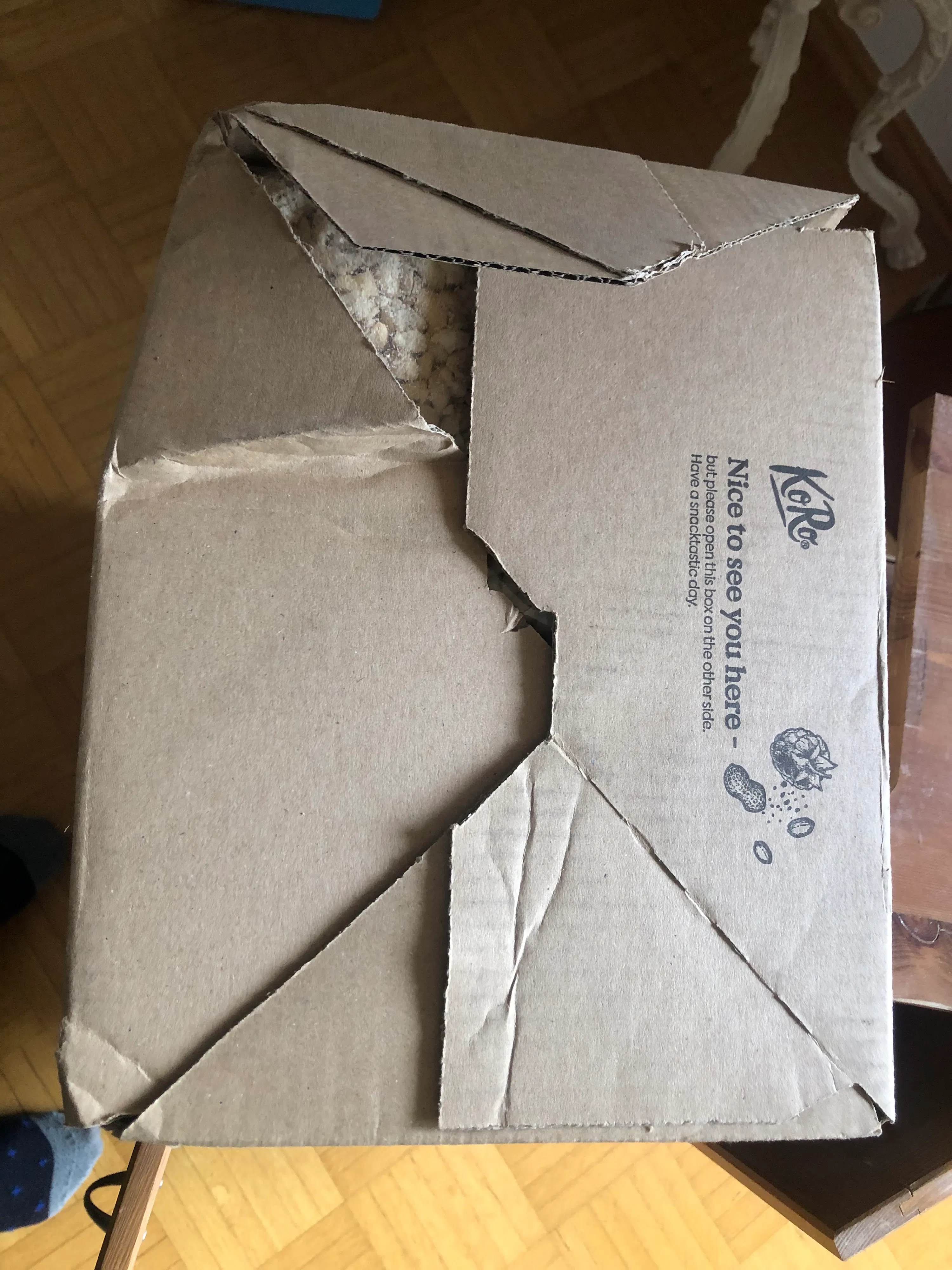 une boîte en carton koro partiellement ouverte, montrant un contenu granuleux clair à l'intérieur, avec le texte "nice to see you here - but please open this box on the other side. have a snacktastic day." imprimé sur le dessus.