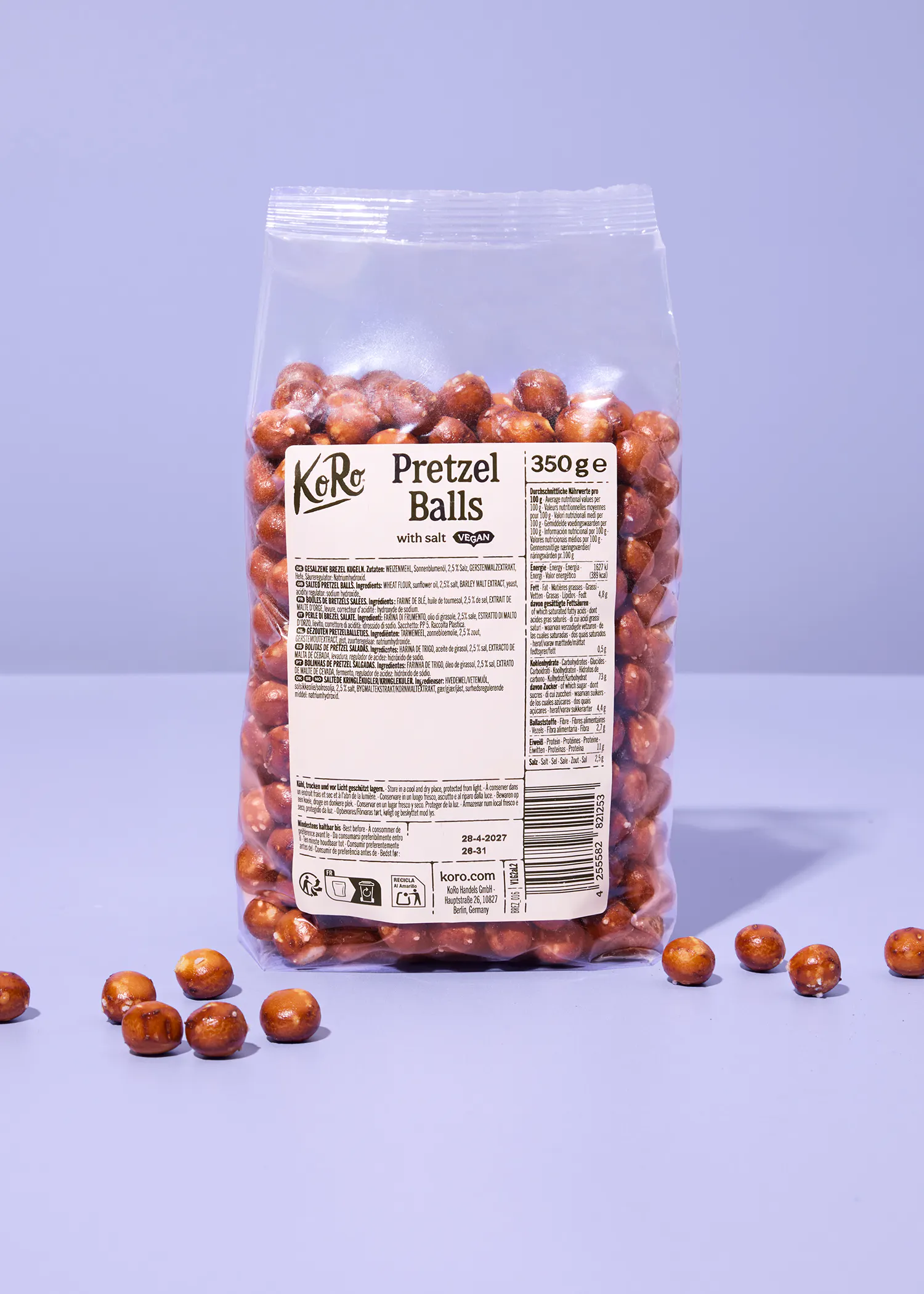 eine durchsichtige tüte mit koro pretzel balls vor einem hellvioletten hintergrund, wobei einige der salzigen teigkugeln lose vor der verpackung liegen.