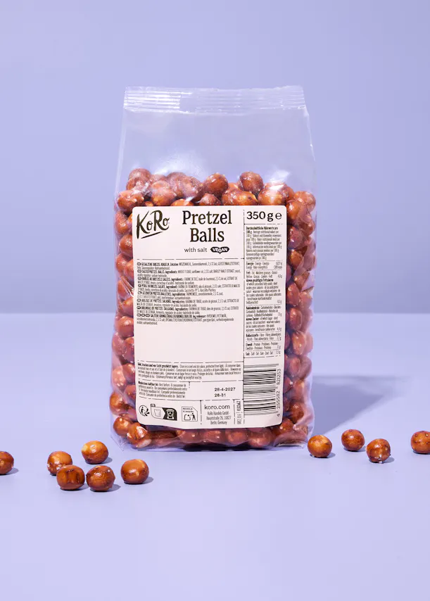 eine durchsichtige tüte mit koro pretzel balls vor einem hellvioletten hintergrund, wobei einige der salzigen teigkugeln lose vor der verpackung liegen.