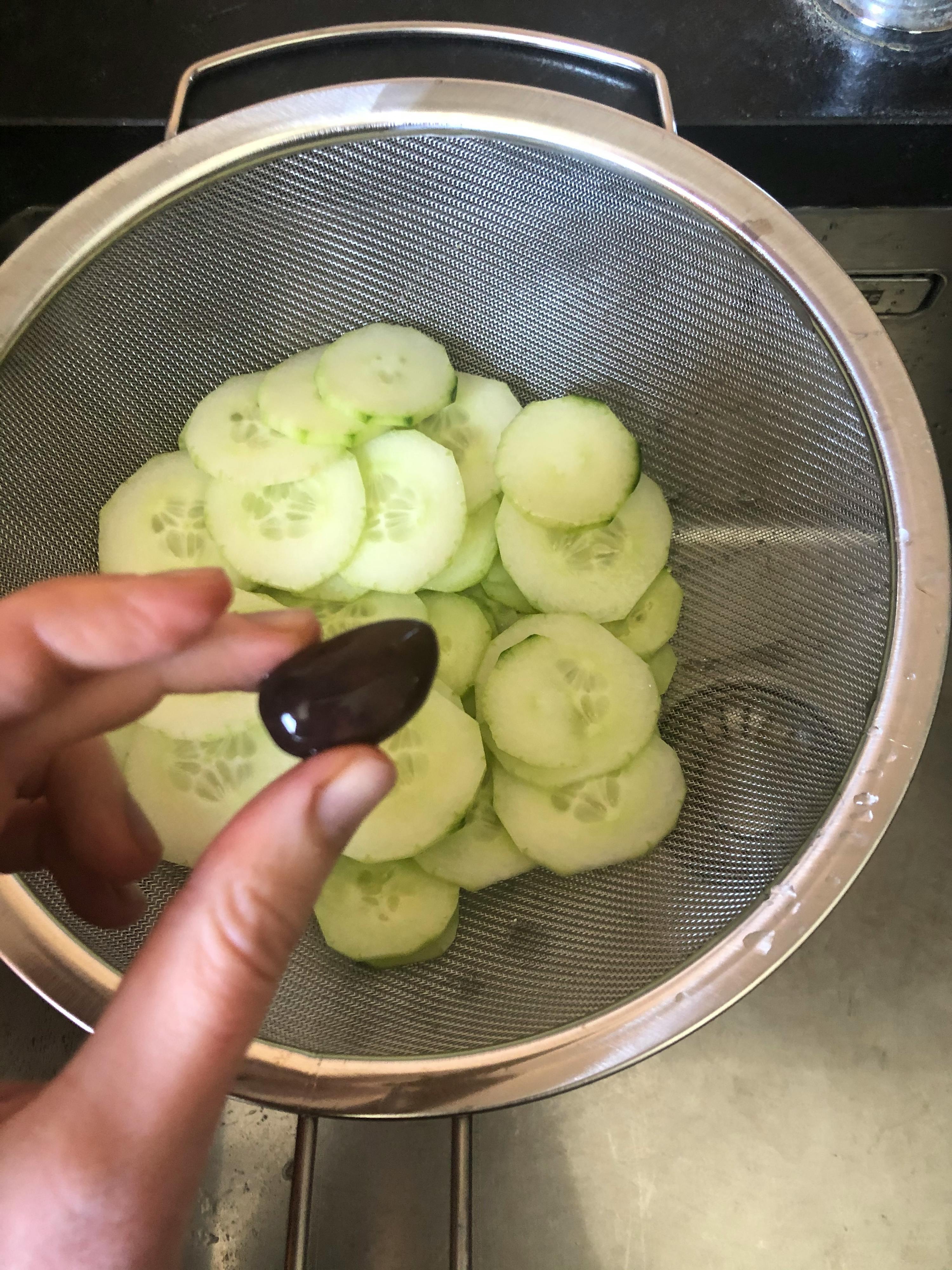 Eine Person hält eine Olive vor einem Strainer mit geschnittenen Gurken