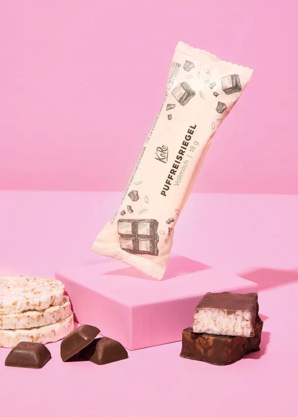 une barre de riz soufflé koro enrobée de chocolat au lait dans son emballage flotte au-dessus d'un bloc rose, entourée de galettes de riz, de morceaux de chocolat et de barres de riz soufflé coupées et entières.