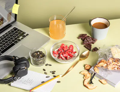 Een bureau met een laptop, een notitieboekje, een koptelefoon, een kom muesli en een kop koffie op een groene achtergrond