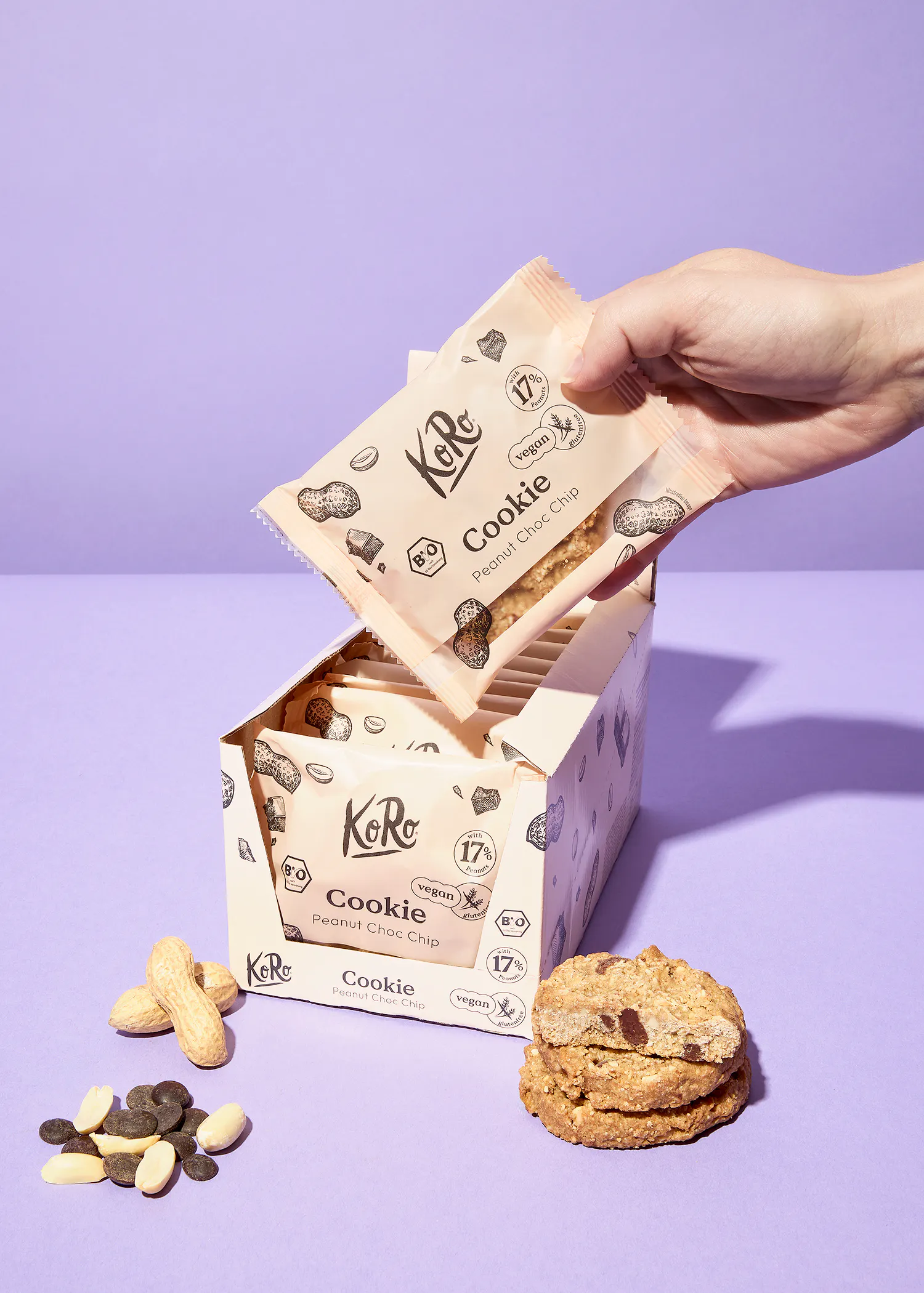 eine hand nimmt eine packung koro cookie peanut choc chip aus einer box, daneben liegen erdnüsse, schokoladenstückchen und zwei kekse auf lila hintergrund.