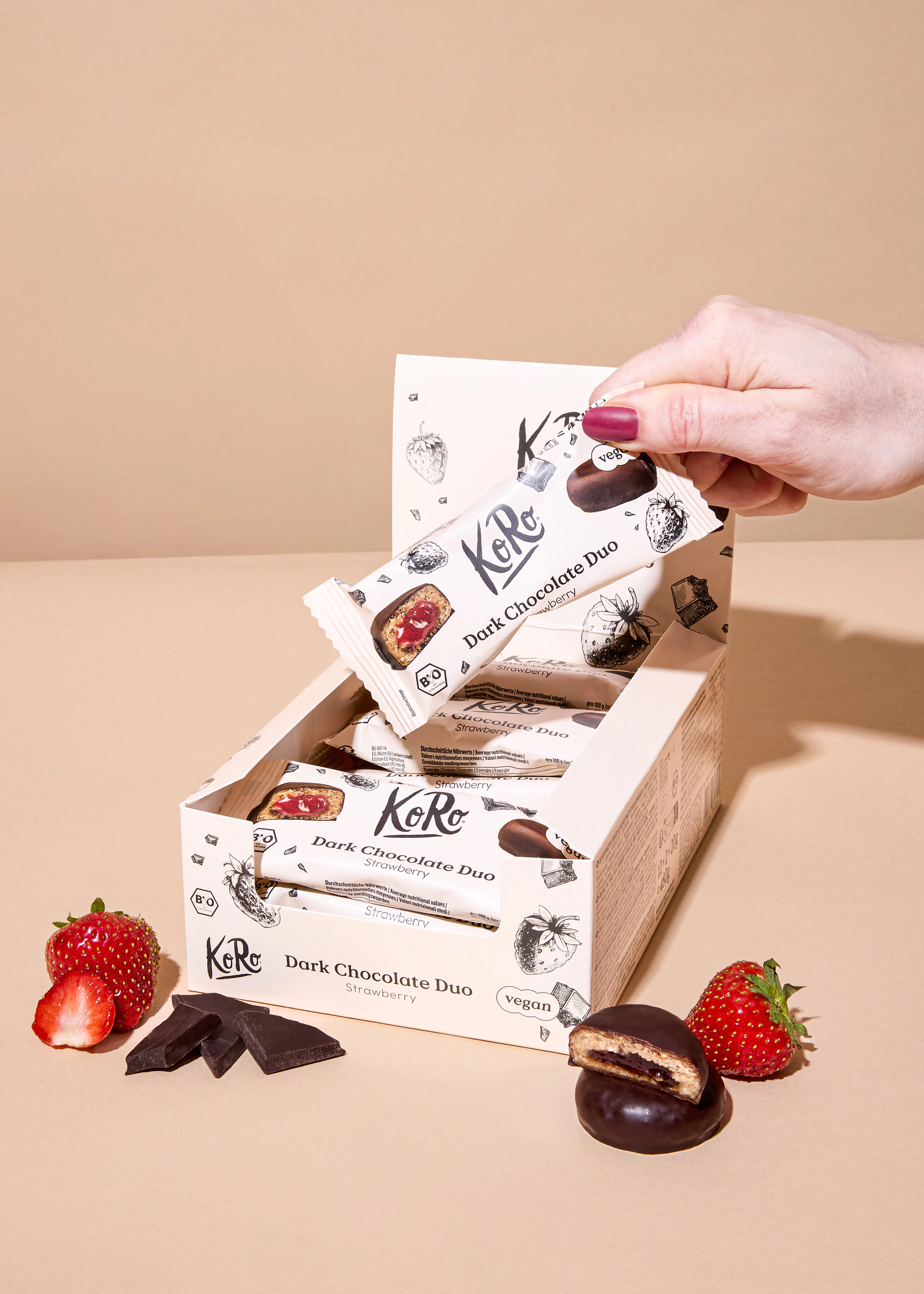 a hand with red nail polish takes a koro dark chocolate duo strawberry bar from a box, surrounded by fresh strawberries and chocolate pieces, with one bar cut open to reveal a strawberry filling.
