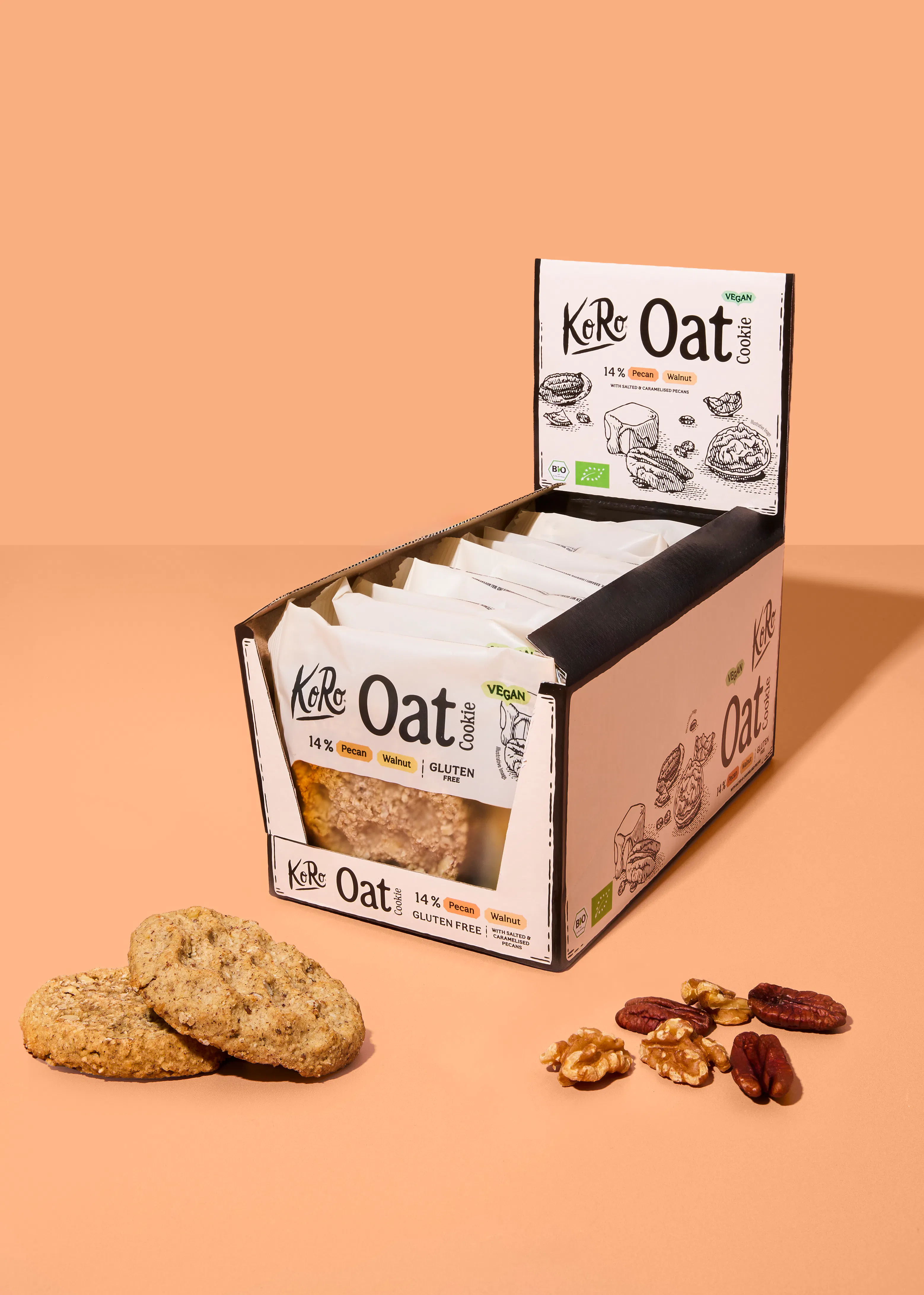 eine verkaufspackung mit koro oat cookies mit pekannuss und walnuss vor einem beigen hintergrund, wobei zwei kekse und einige nüsse davor liegen.