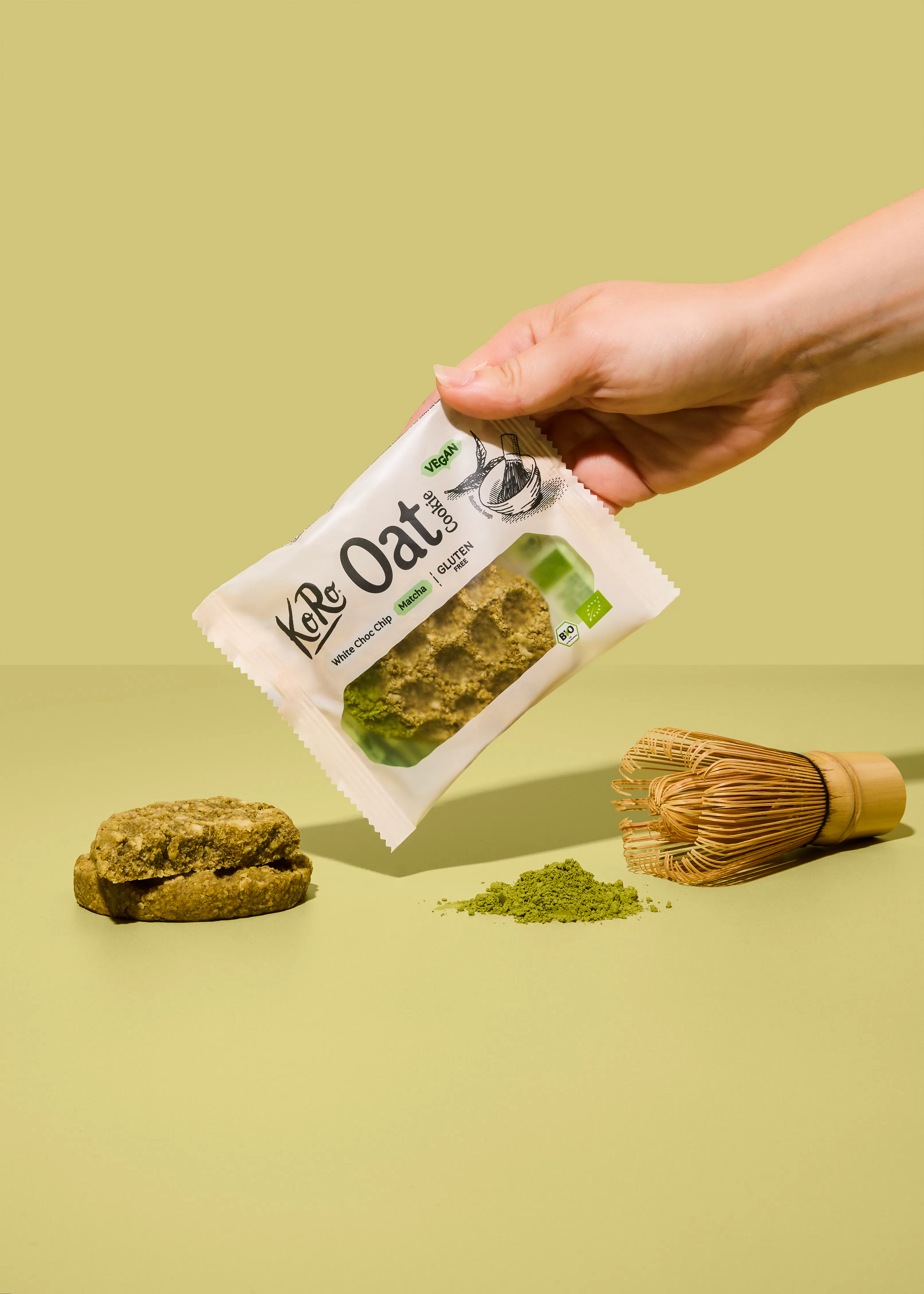 eine hand hält eine verpackung eines koro matcha white choc chip cookies über einem tisch, auf dem ein weiterer keks, etwas matchapulver und ein bambusbesen liegen.