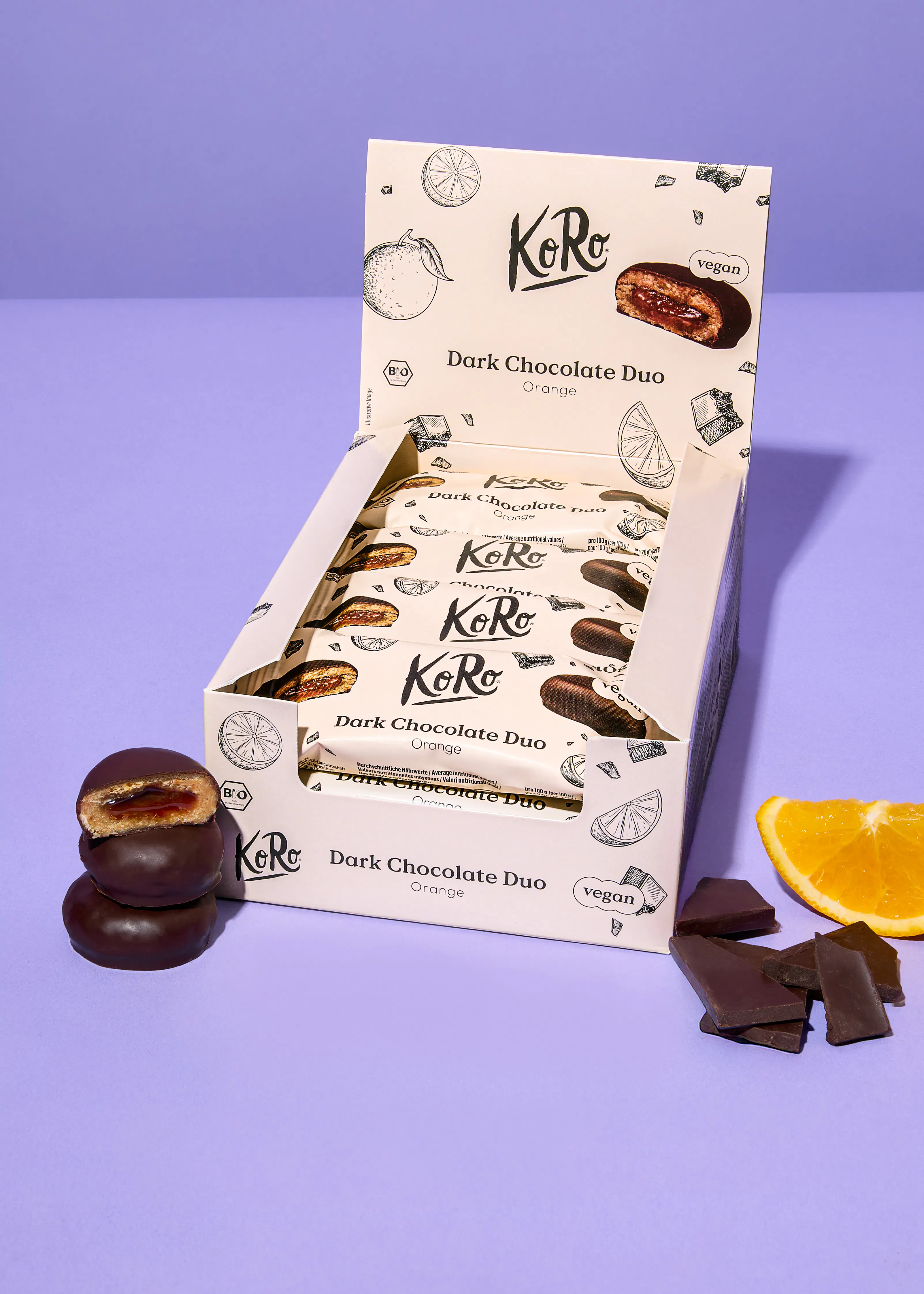 eine weiße displaybox mit koro dark chocolate duo orange riegeln, von denen einer und eine daneben liegende schokoladenscheibe eine orangefarbene füllung zeigen, ergänzt durch eine orangenscheibe und schokoladenstücke auf lila hintergrund.