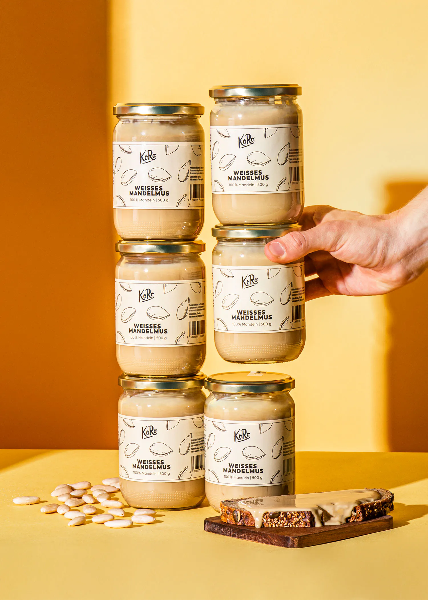 une main empile des pots de beurre d'amande blanc koro sur une table jaune, avec des amandes éparpillées et une tranche de pain tartinée de beurre d'amande.