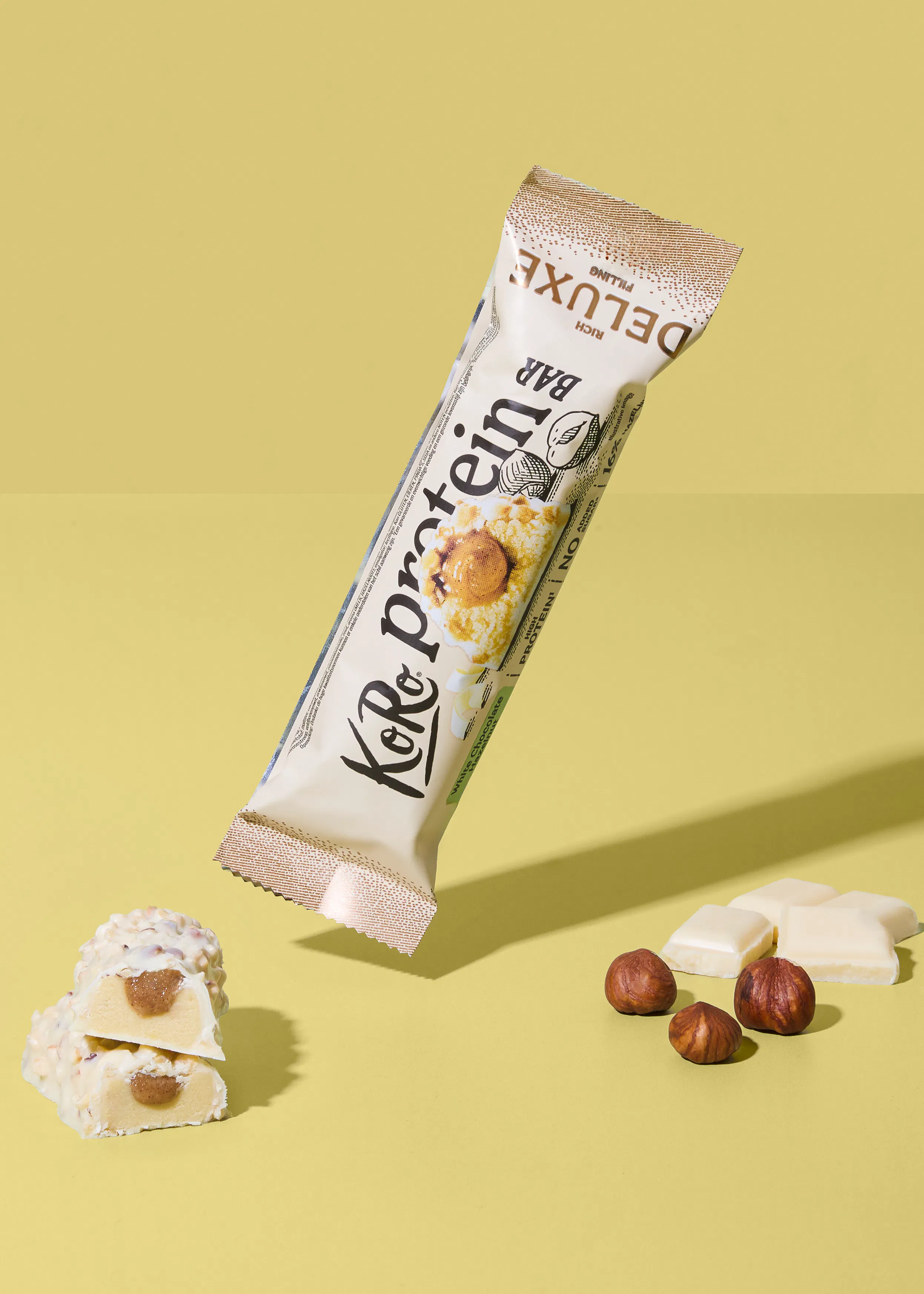 une barre protéinée kor&protein deluxe à la garniture riche, ouverte pour révéler son intérieur, est présentée avec une barre coupée montrant une garniture au caramel, des noisettes et des morceaux de chocolat blanc sur un fond jaune.