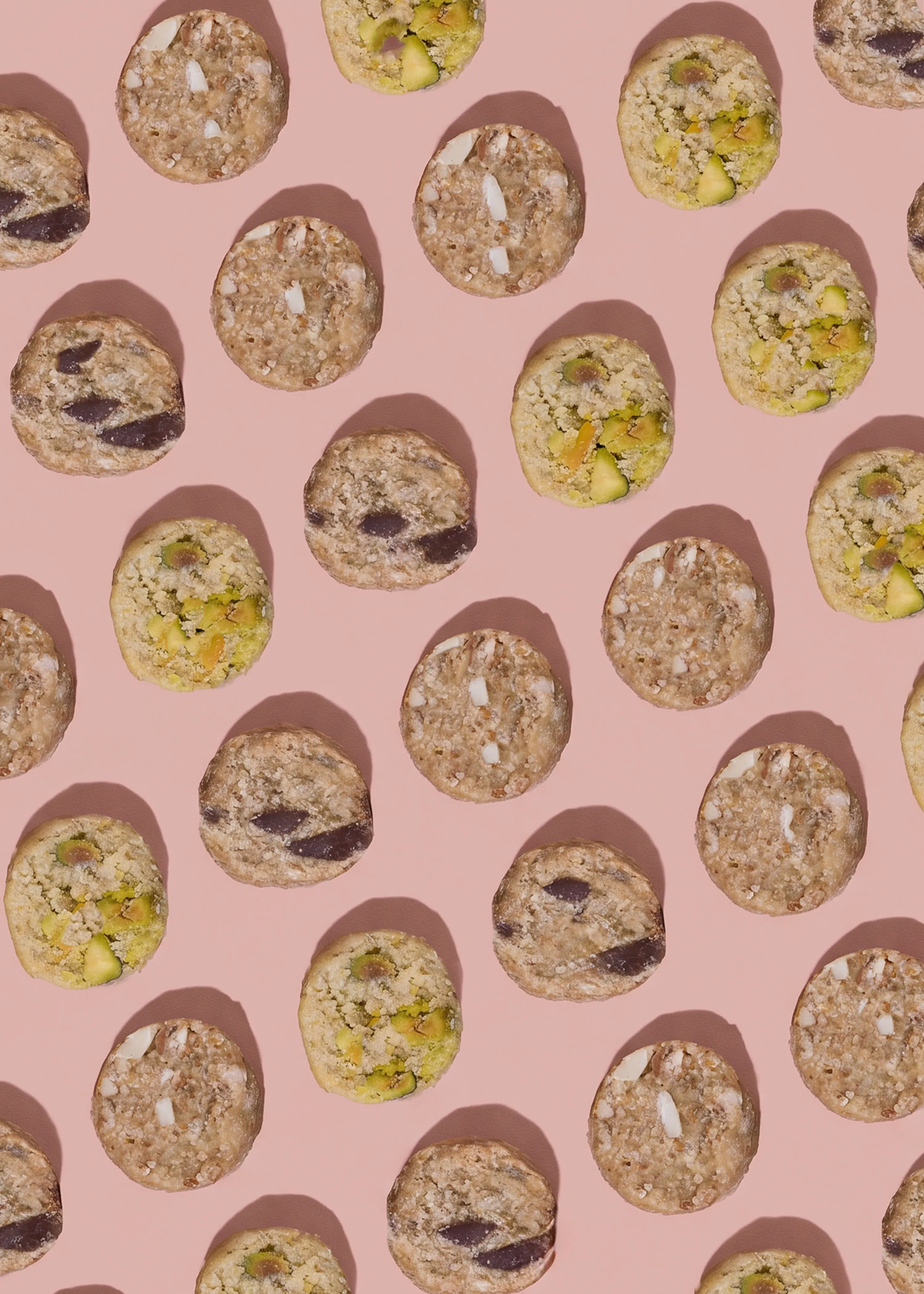 un motivo ripetuto di biscotti rotondi di tre tipi: con gocce di cioccolato, con pistacchi e con mandorle, su uno sfondo rosa.