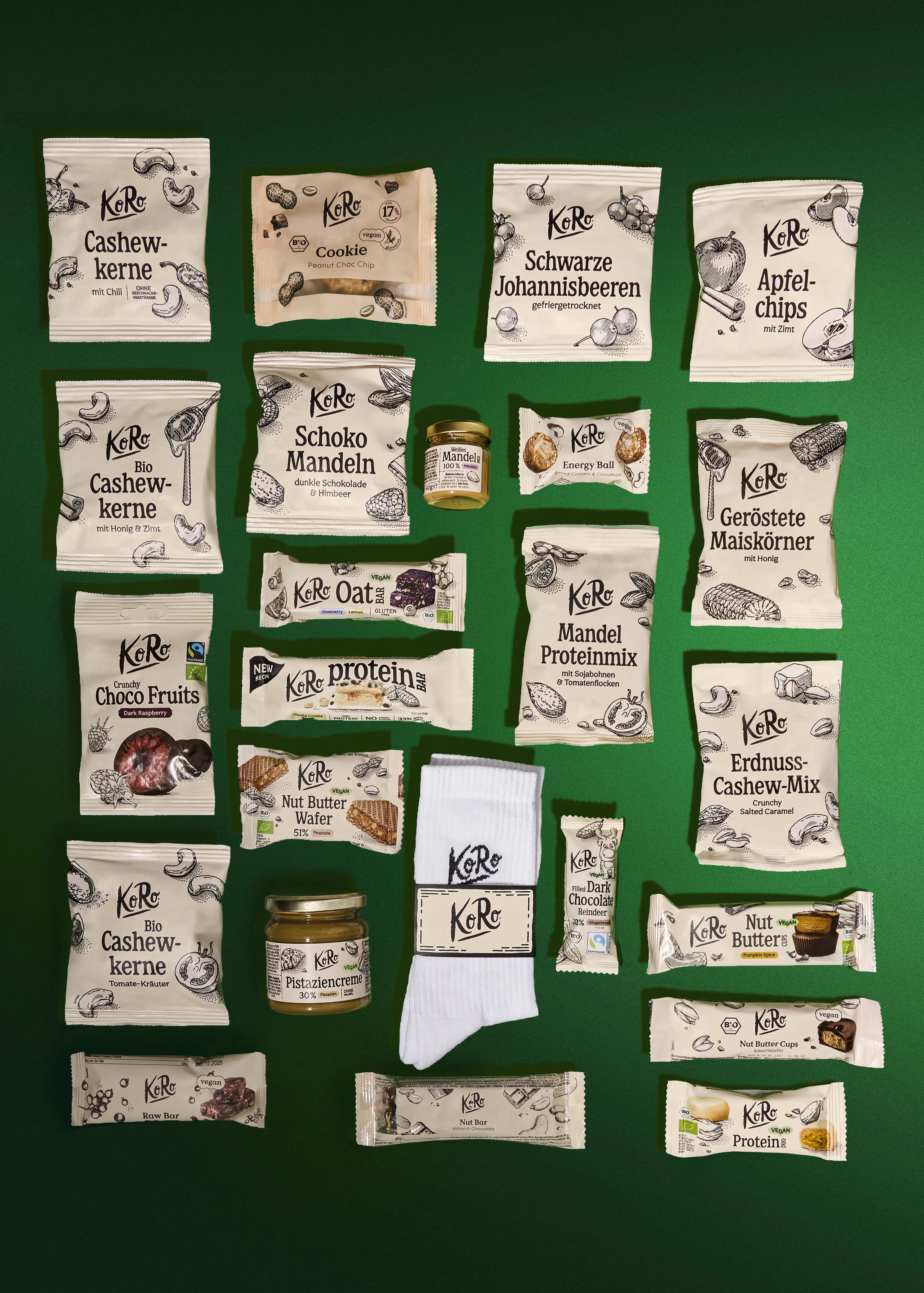 eine flache draufsicht auf eine vielzahl von koro-lebensmittelprodukten, darunter verschiedene nussmischungen, riegel, aufstriche und ein paar weiße socken, die auf einem dunkelgrünen hintergrund angeordnet sind.