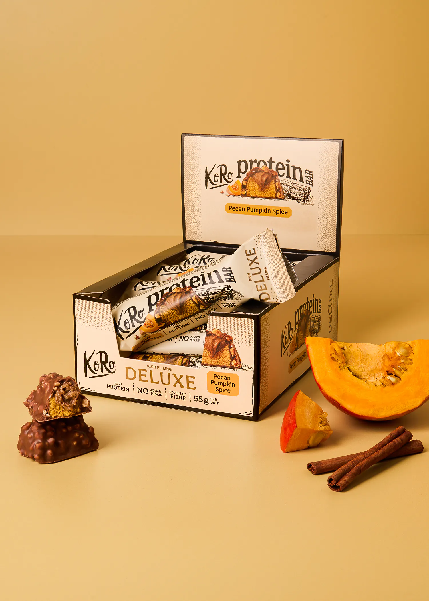 eine geöffnete verkaufsbox mit koro proteinriegeln der sorte pecan pumpkin spice, daneben liegen ein stück kürbis und zimtstangen sowie ein halbierter riegel.