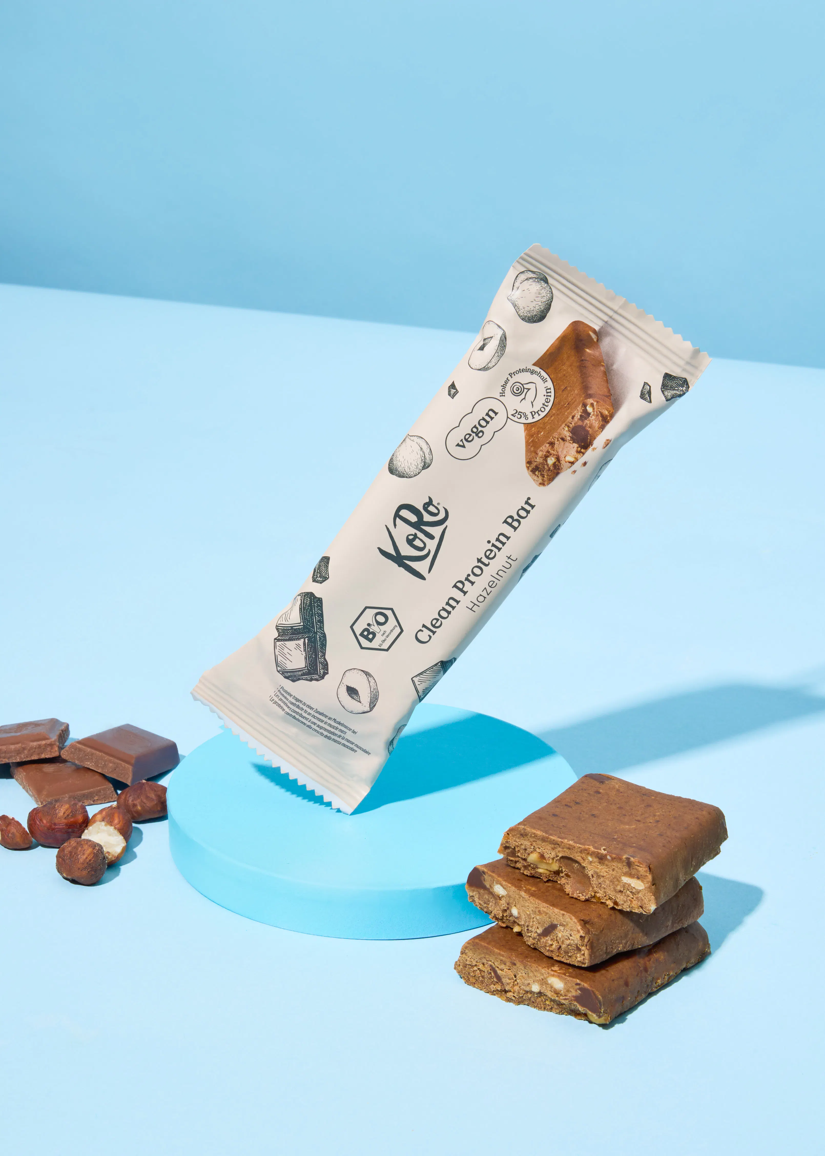 une barre protéinée koro clean saveur noisette dans son emballage ouvert, montrant l'intérieur avec des noisettes, et des morceaux coupés de la barre empilés à côté, avec des noisettes et des carrés de chocolat sur un fond bleu.