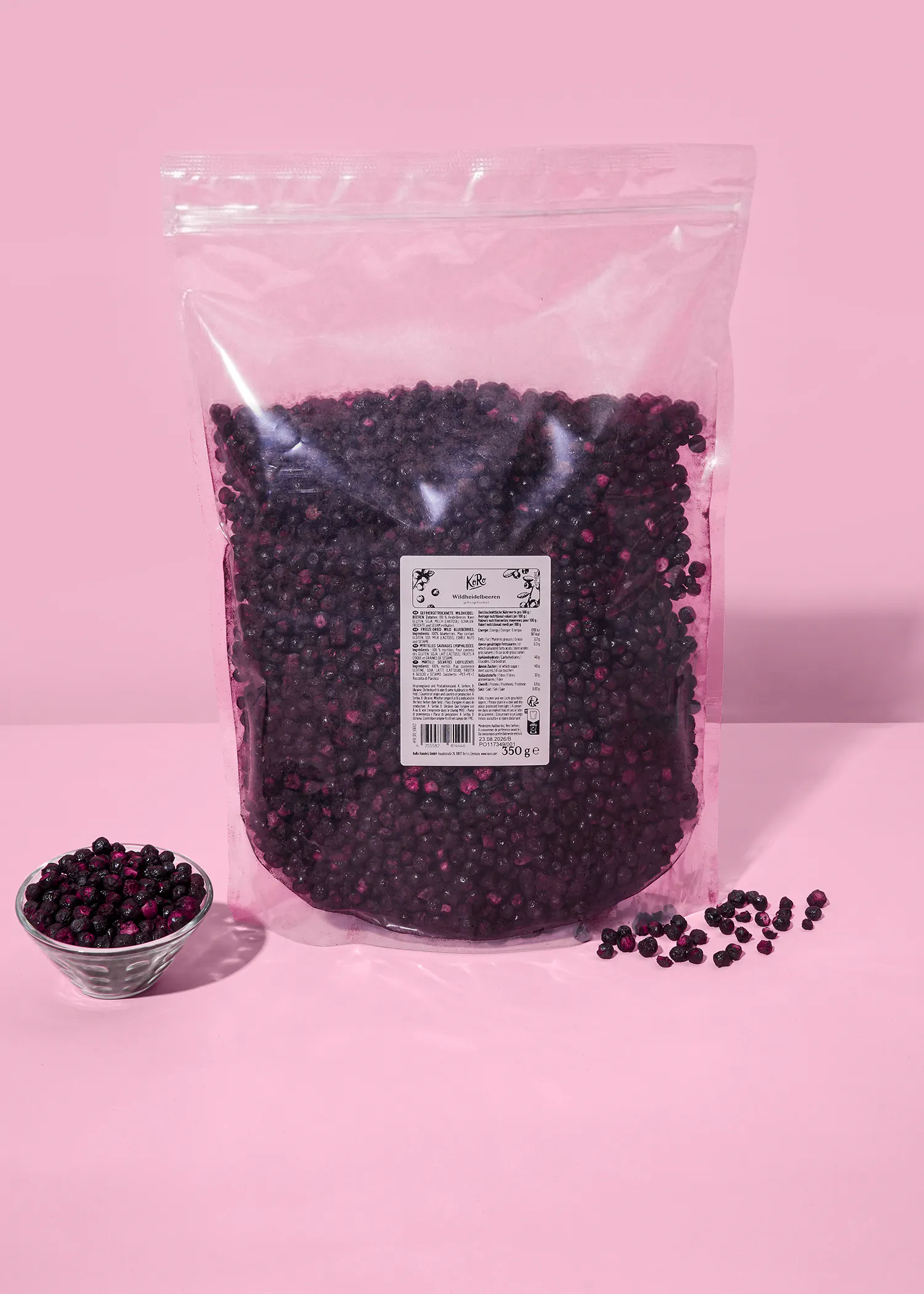 una bolsa grande de arándanos silvestres con una etiqueta de 350 gramos, un tazón pequeño de arándanos y algunos arándanos esparcidos sobre una superficie rosa.