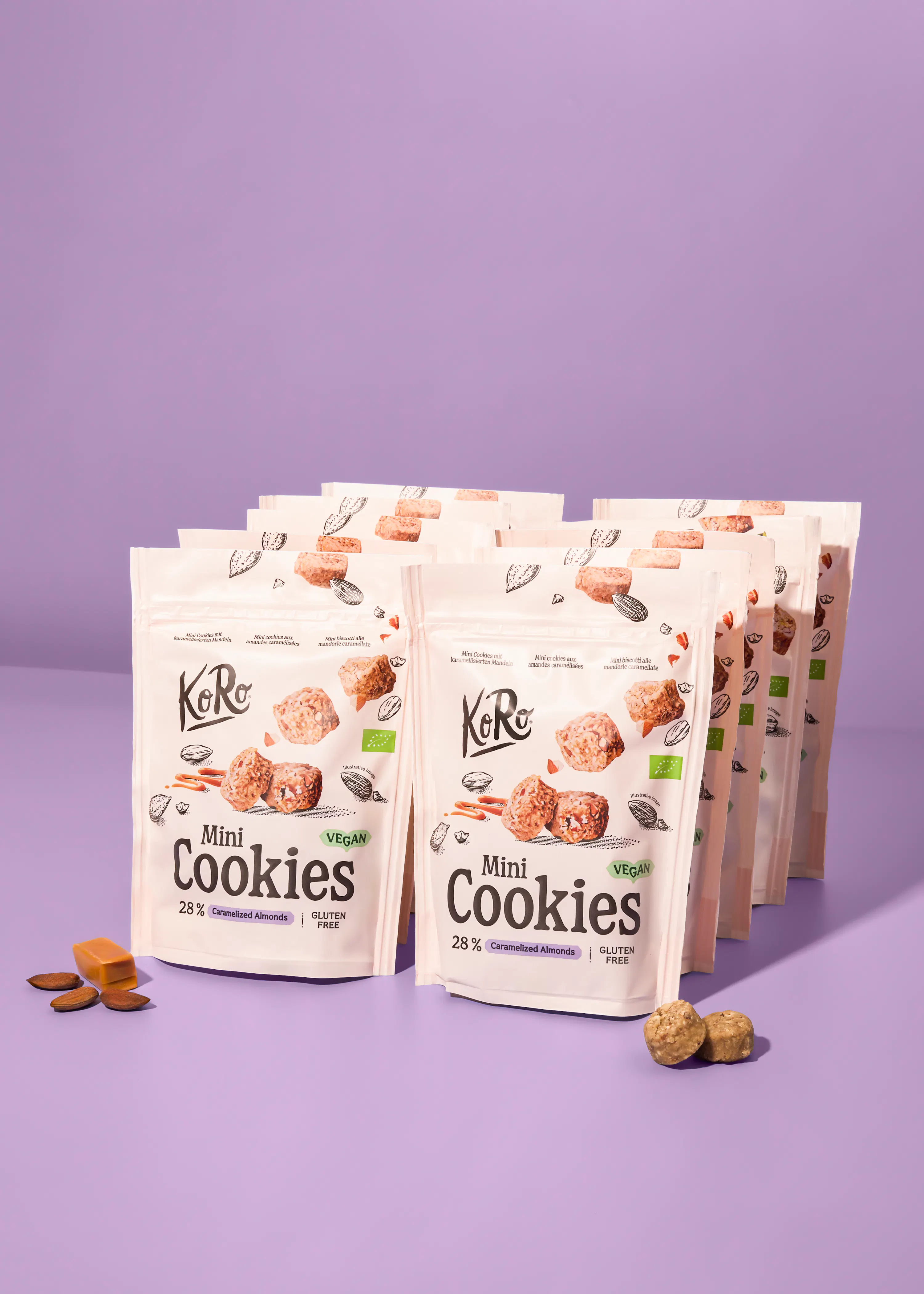 mehrere packungen koro mini cookies mit karamellisierten mandeln und glutenfrei-etikett, zusammen mit losen mandeln und karamellstücken auf lila hintergrund.