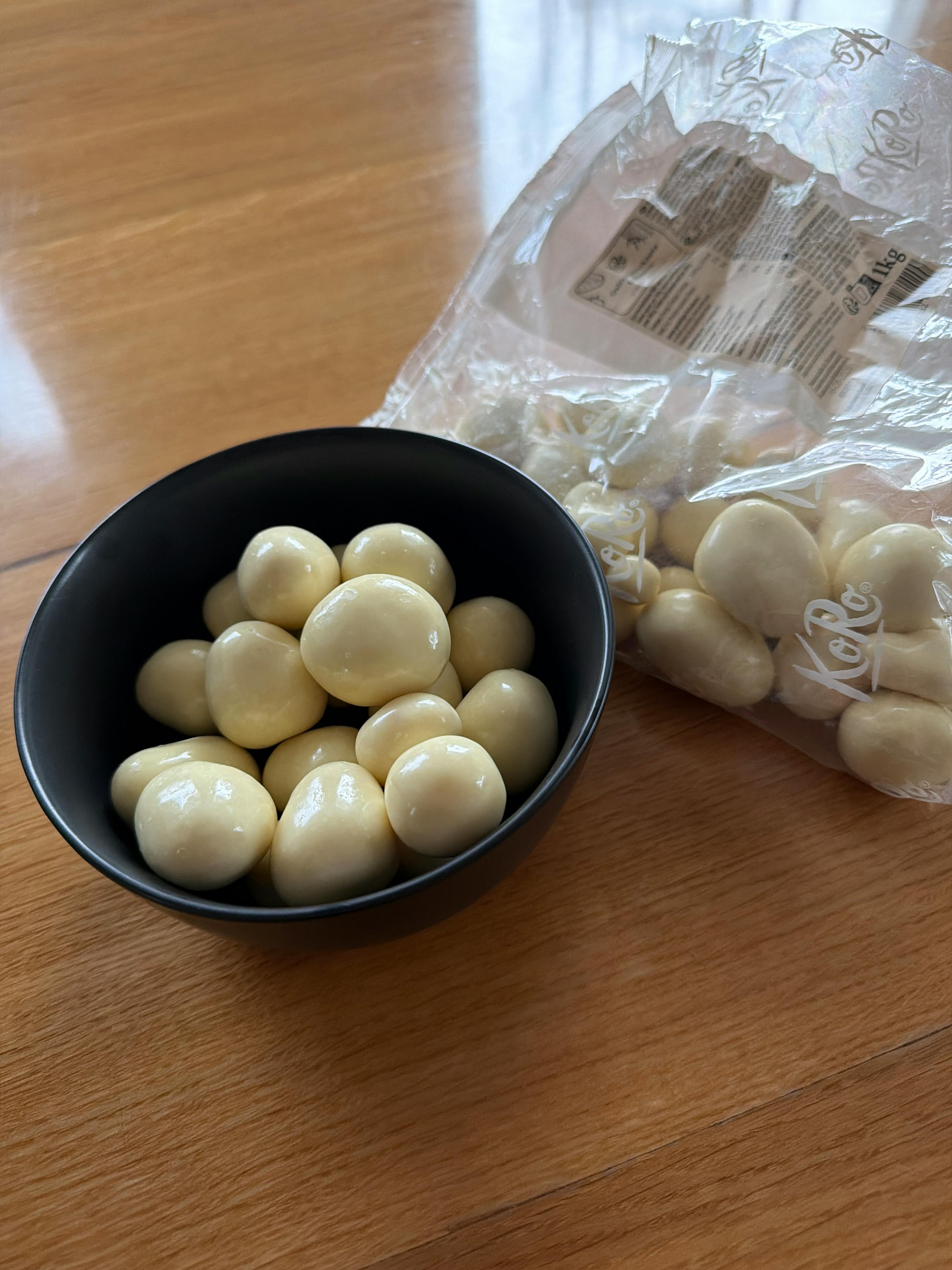 una ciotola nera piena di caramelle bianche rotonde e un sacchetto di plastica delle stesse caramelle su una superficie di legno.
