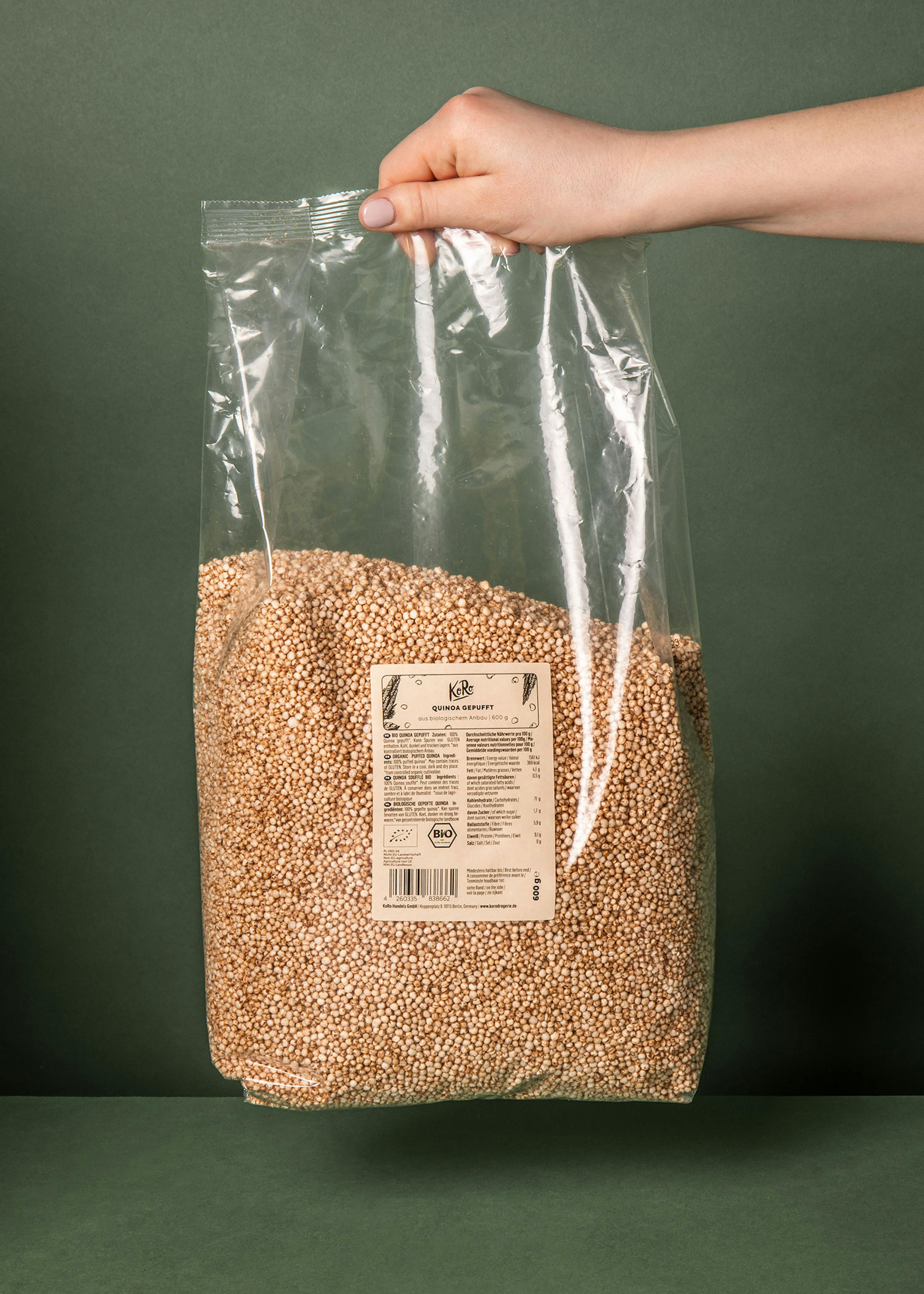 eine hand hält einen großen, durchsichtigen plastikbeutel mit gepufftem quinoa und einem etikett mit der aufschrift 'kore quinoa gepufft' vor einem dunkelgrünen hintergrund.