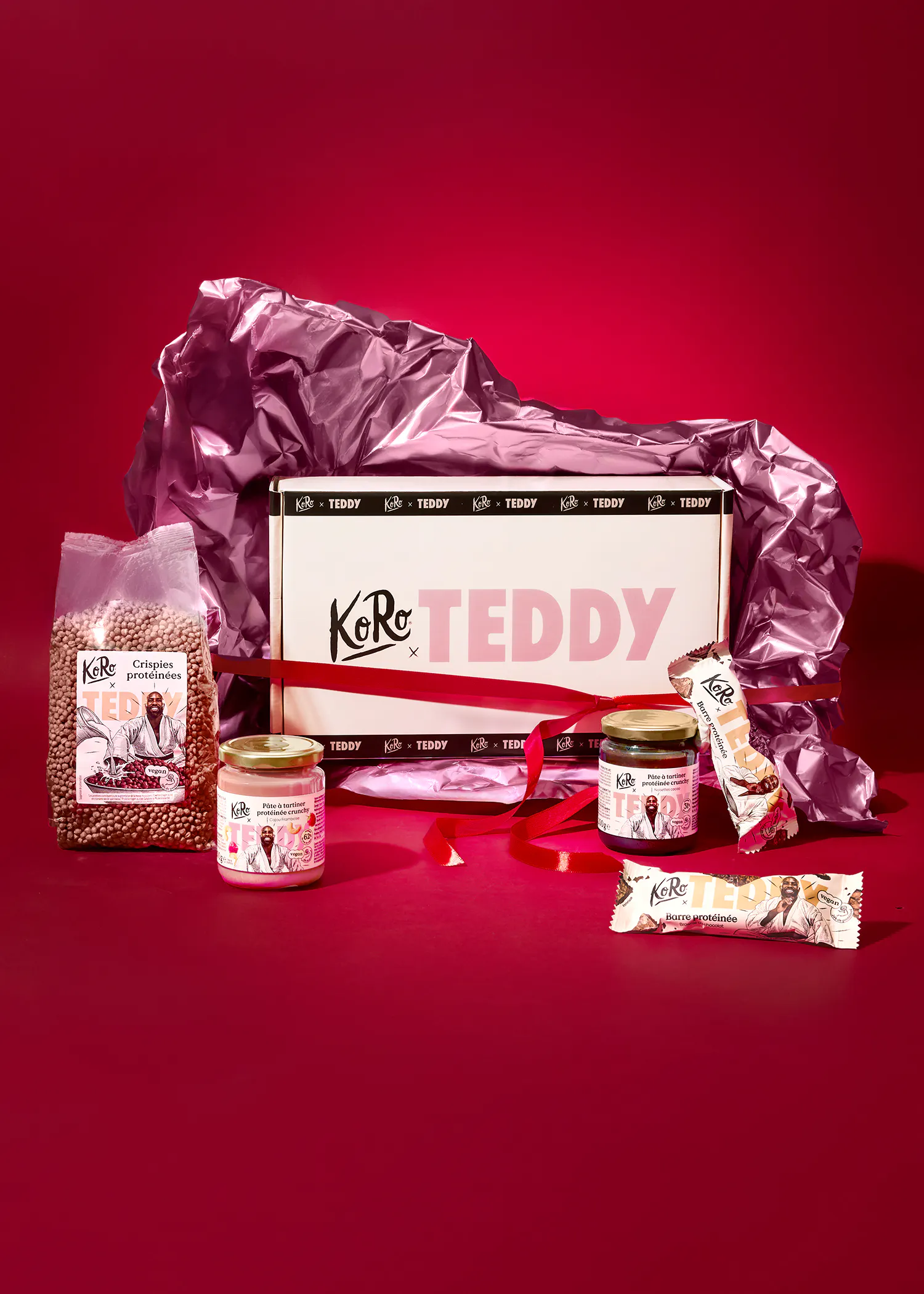 une boîte cadeau koro x teddy ouverte, remplie de papier d'emballage rose, présente des produits protéinés comme des croustilles, des pâtes à tartiner et des barres, sur un fond rouge.