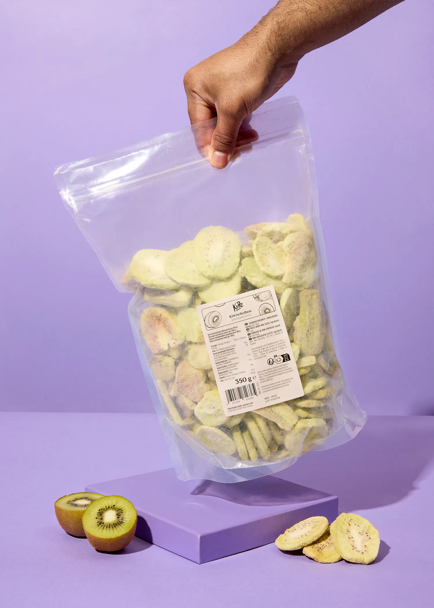 a hand holds a clear bag of dried kiwi slices above fresh kiwi halves and more dried slices on a purple surface.