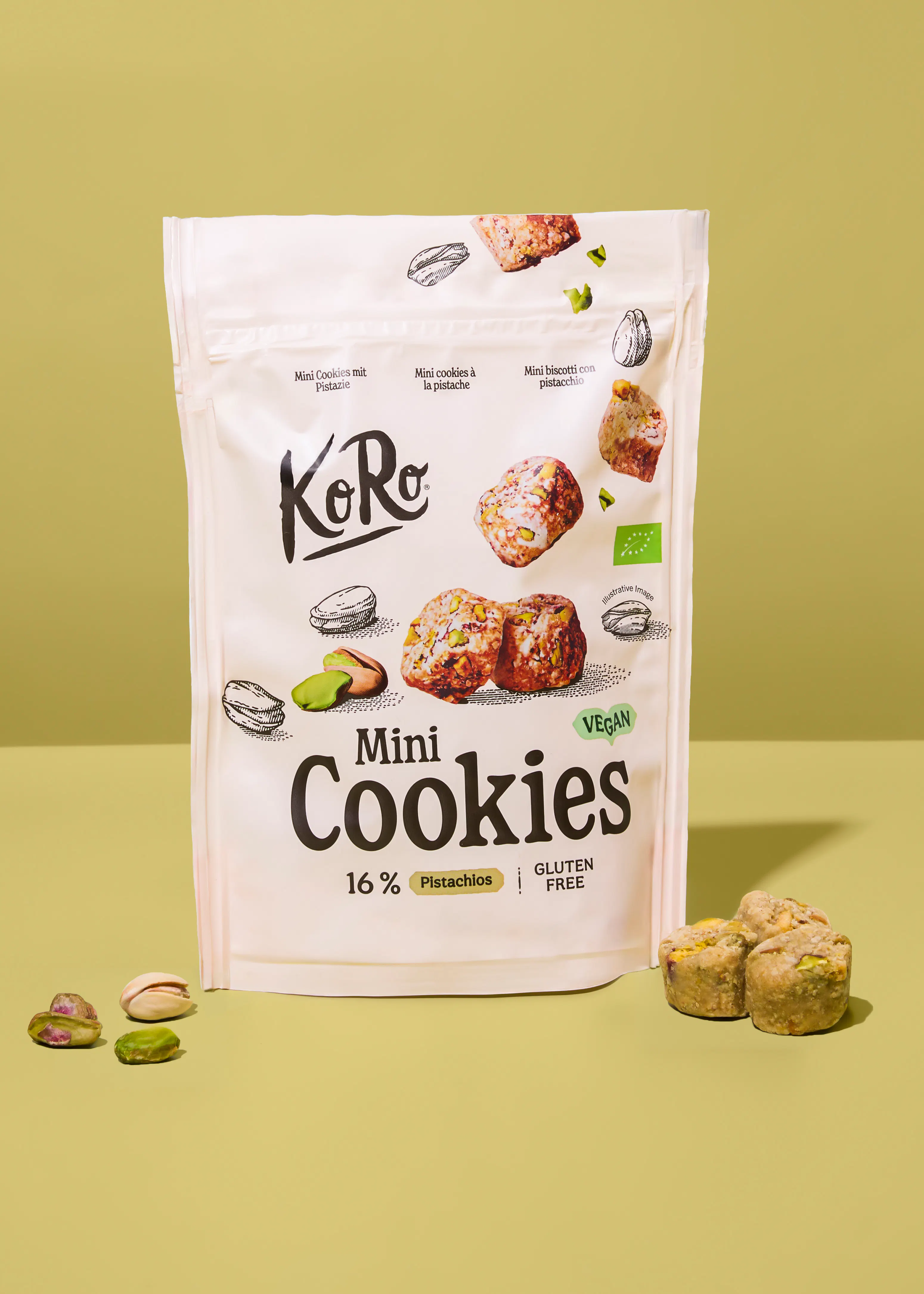 un paquete blanco de mini galletas koro con pistachos, etiquetado como vegano y sin gluten, se muestra sobre una superficie verde con galletas y pistachos esparcidos.