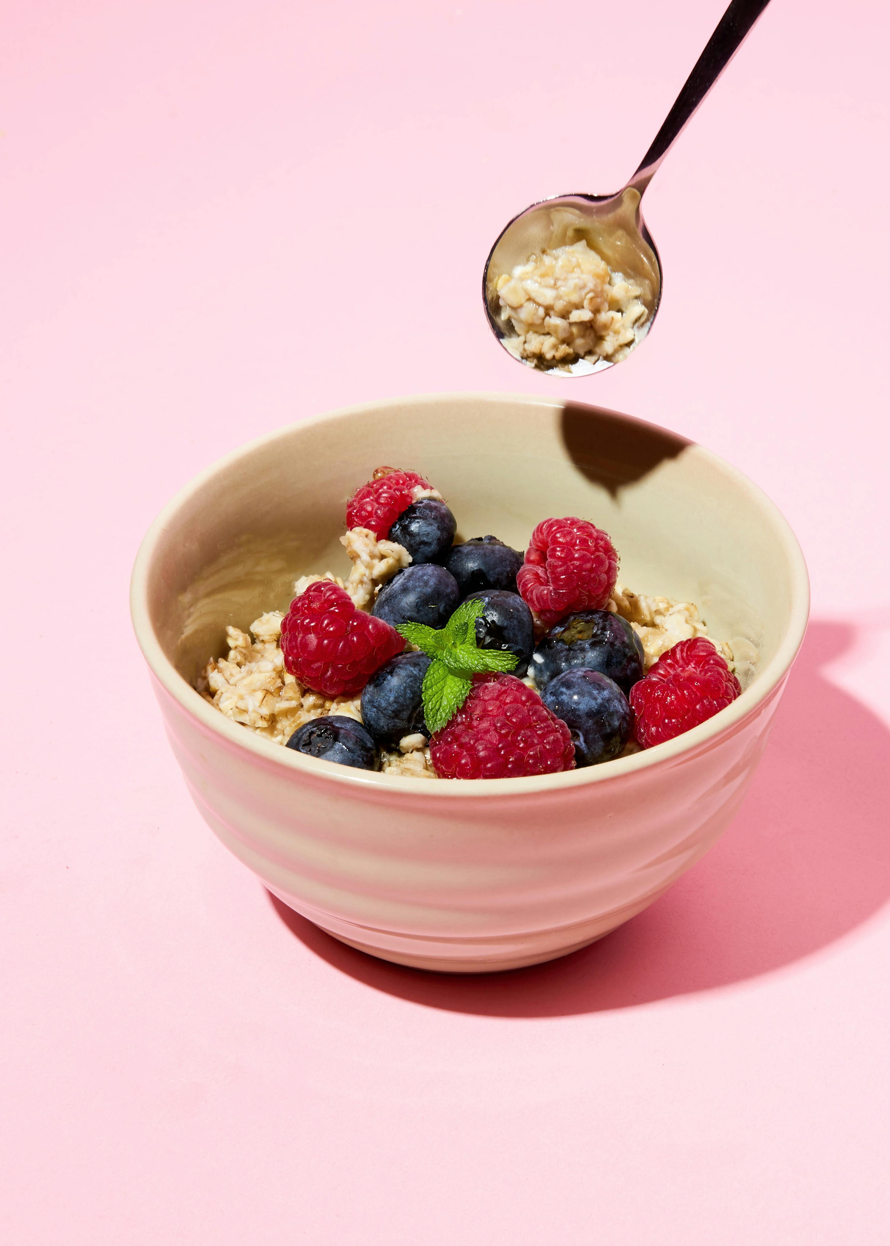 a bowl of oatmeal topped with raspberries, blueberries, and a mint sprig, with a spoon holding more oatmeal hovering above, all on a pink background.
