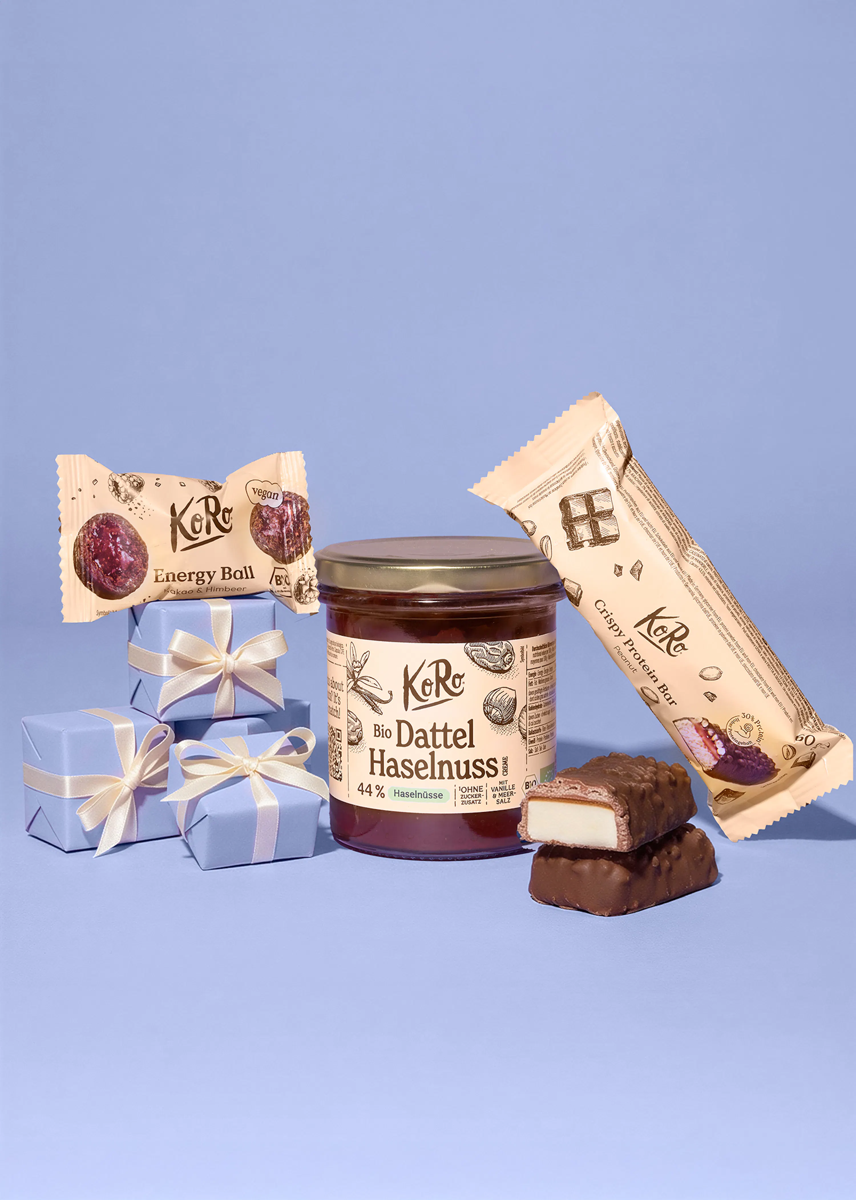 eine auswahl an koro-produkten, darunter vegane energy balls, ein bio-dattel-haselnuss-aufstrich, ein crispy protein bar und zwei schokoriegel, zusammen mit drei kleinen hellblauen geschenkboxen auf hellblauem hintergrund.