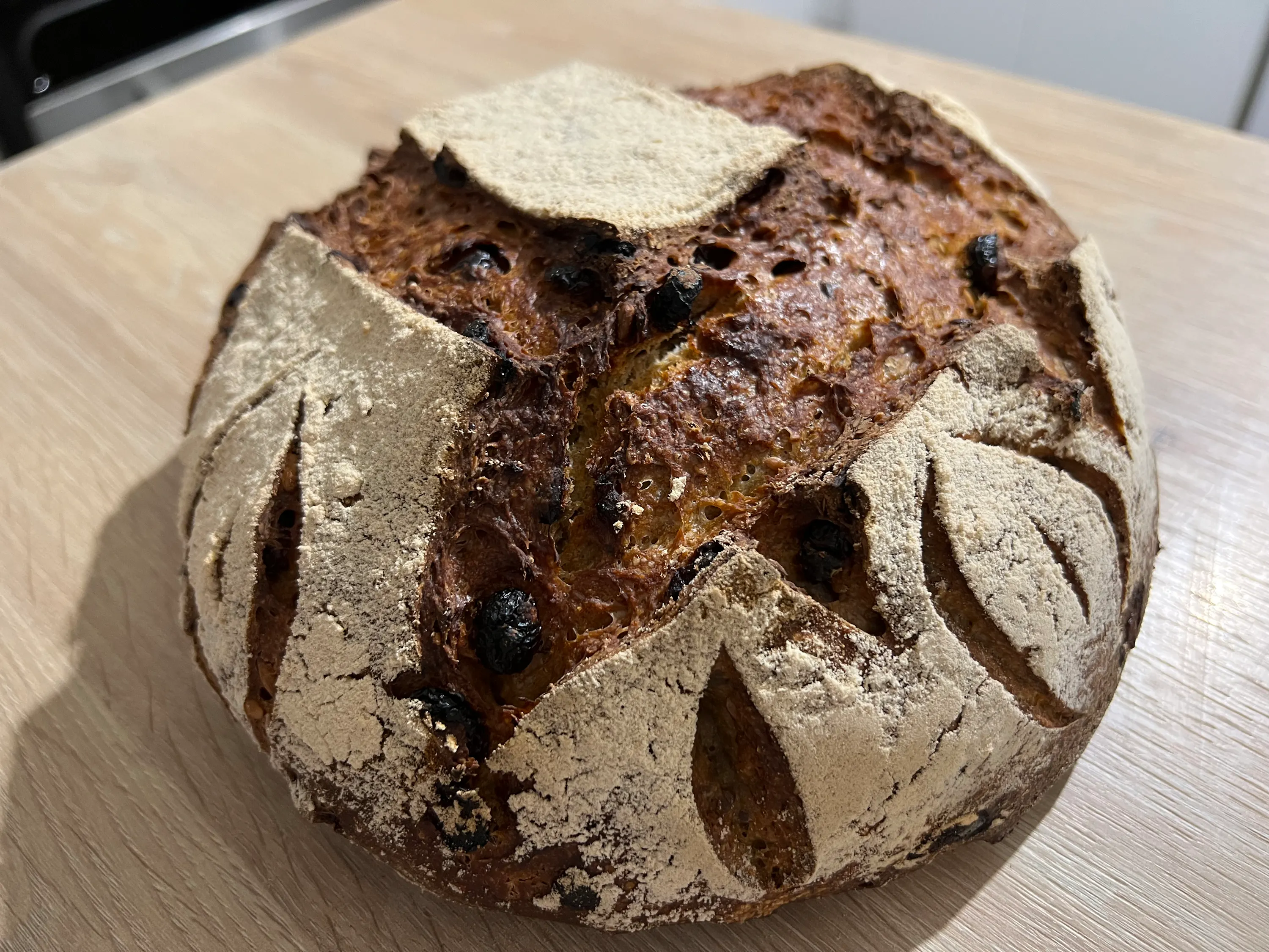 ein rundes, dunkles sauerteigbrot mit rosinen und mehlierten schnitten auf einer hellen holzoberfläche.