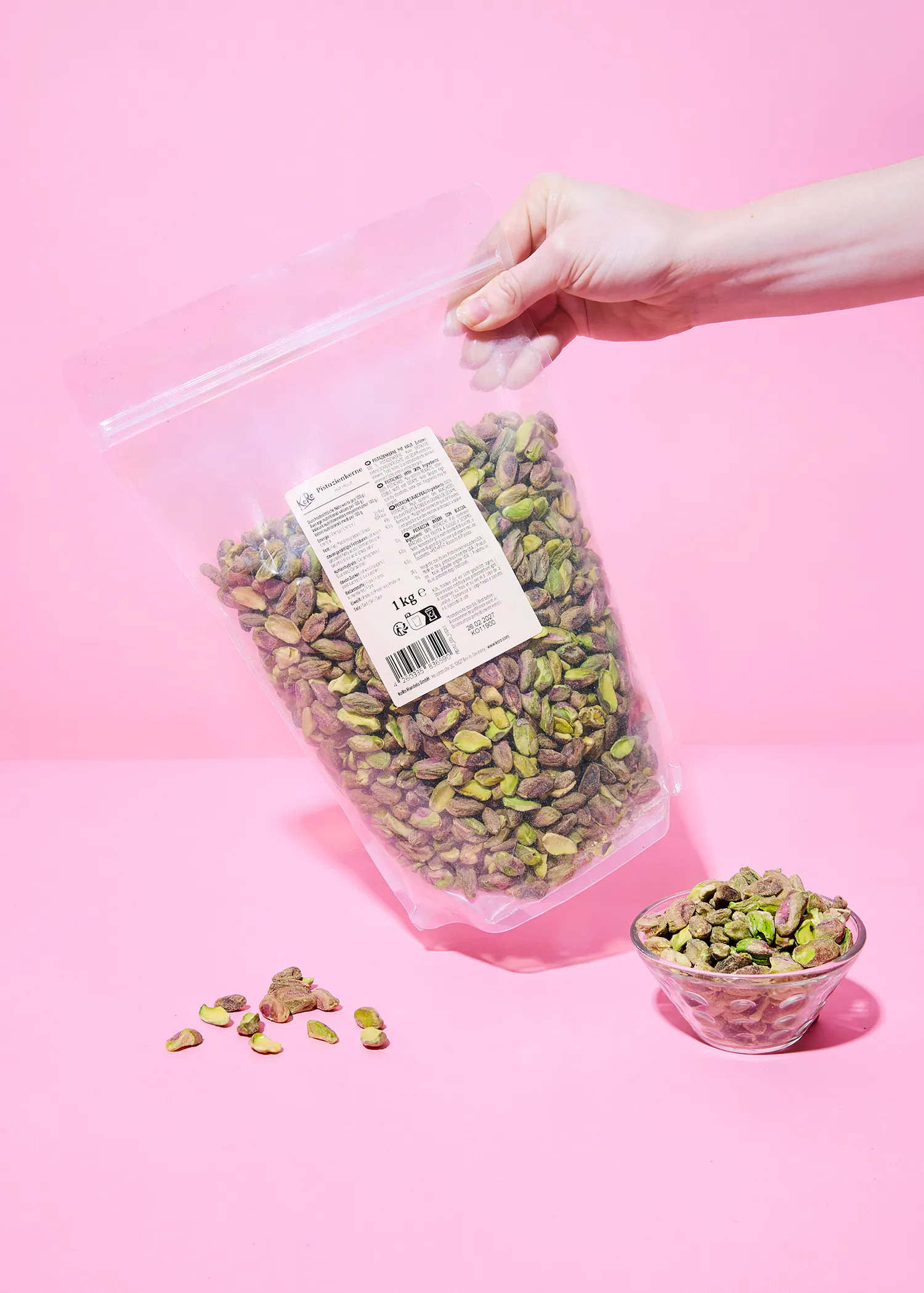 une main tient un sac transparent d'un kilogramme de pistaches décortiquées au-dessus d'un petit bol de pistaches et de quelques pistaches éparpillées sur un fond rose.