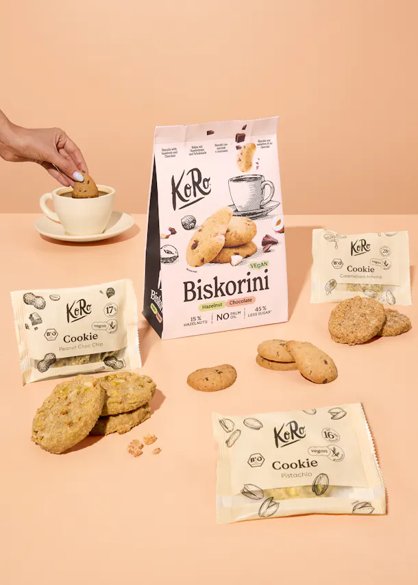 eine hand taucht einen keks in eine tasse, umgeben von verschiedenen KoRo-keksen und -verpackungen, darunter biskorini haselnuss-schokolade.