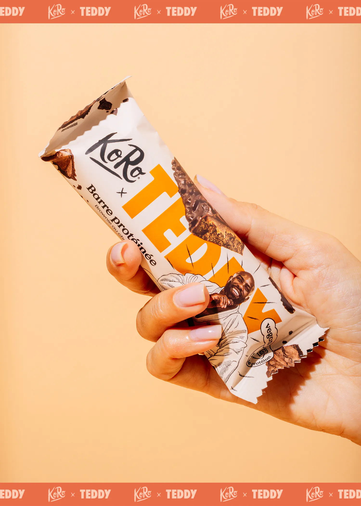 une main tient une barre protéinée koro x teddy saveur brownie au chocolat, avec un homme souriant sur l'emballage, sur un fond orange clair.
