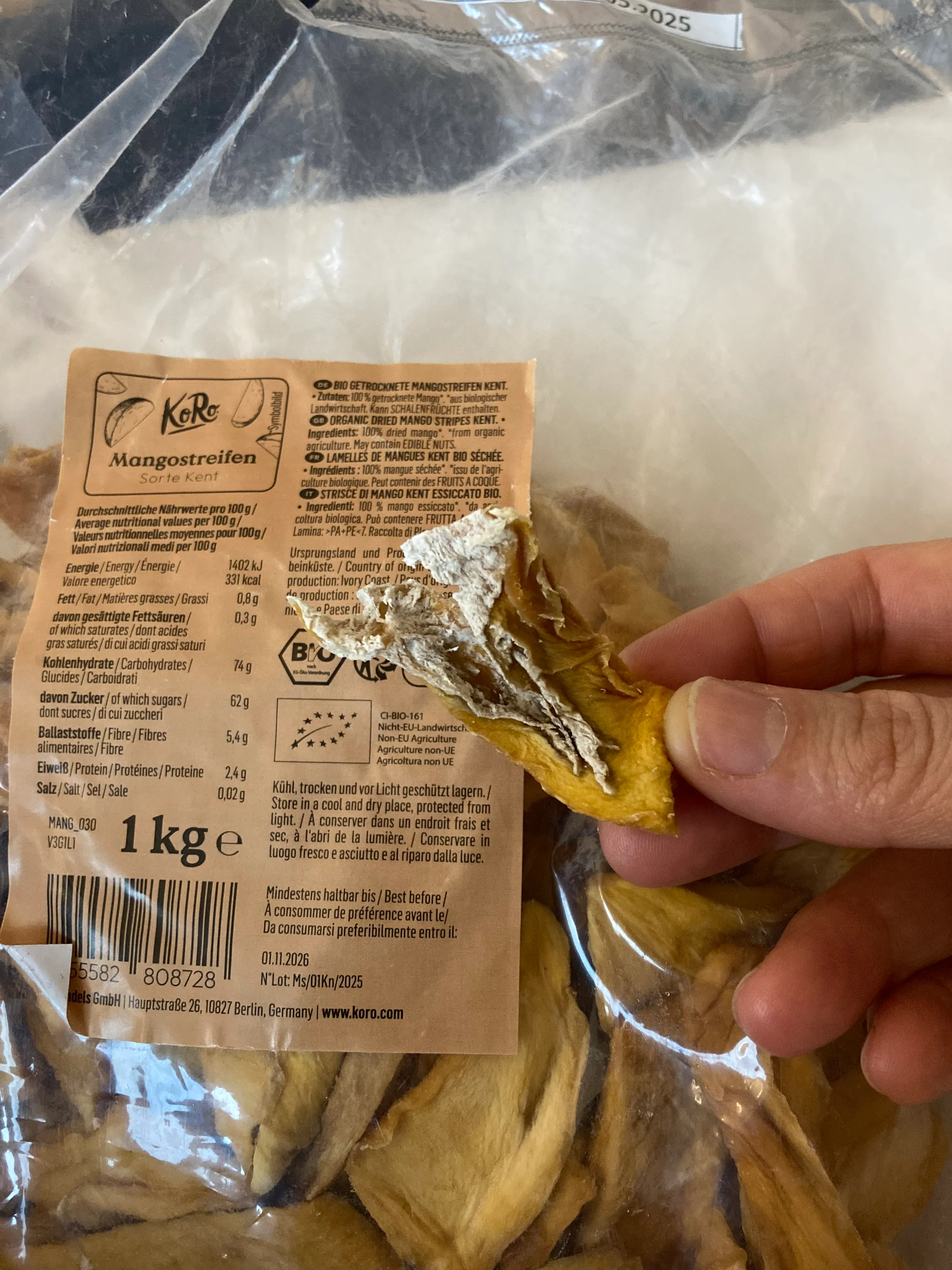 une main tient un morceau de mangue séchée moisi provenant d'un sachet transparent avec une étiquette de produit.