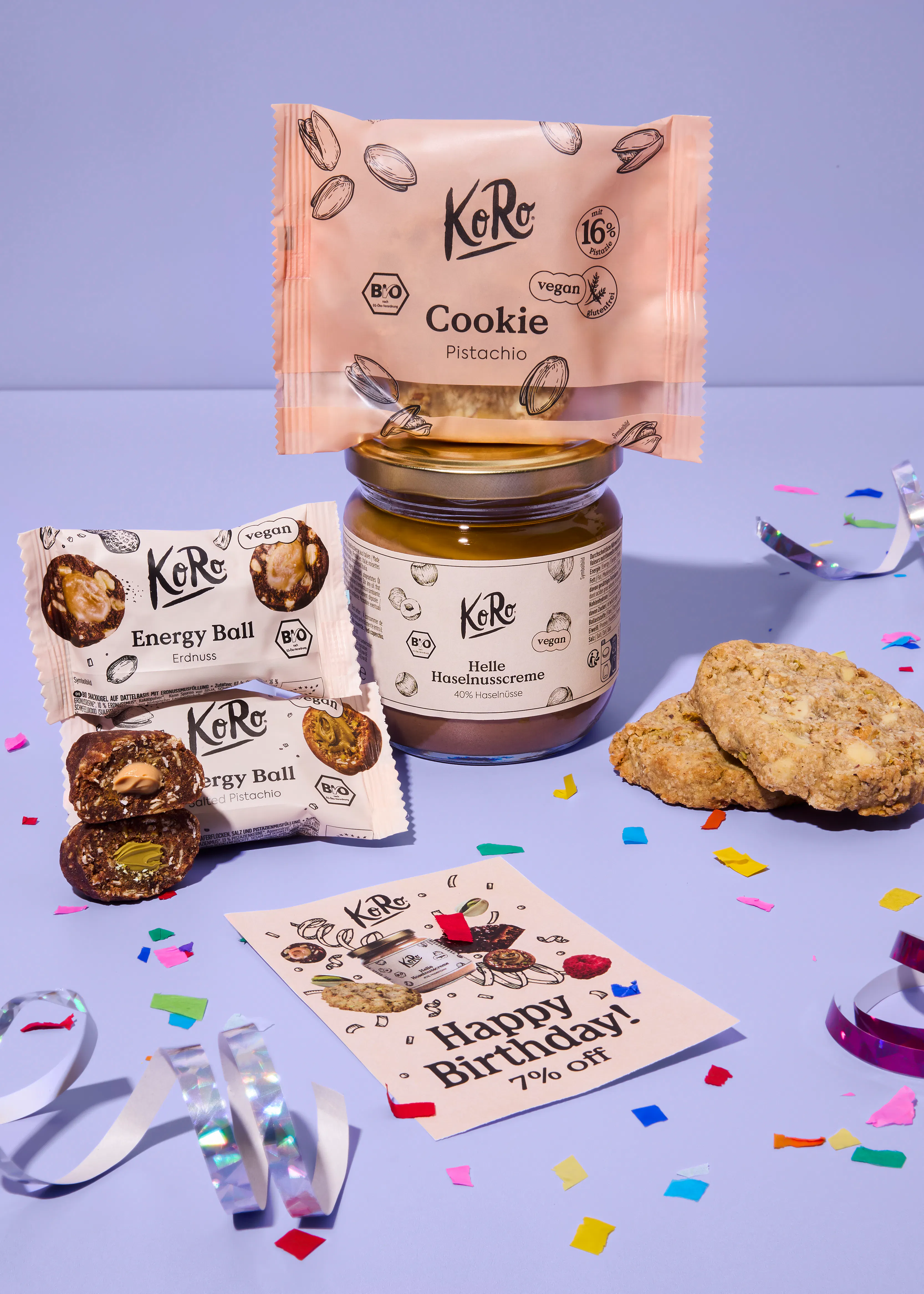 eine anordnung von koro-produkten bestehend aus einem pistazien-cookie, einem glas helle haselnusscreme, zwei energy balls und einer geburtstagskarte auf einem lila hintergrund mit konfetti.