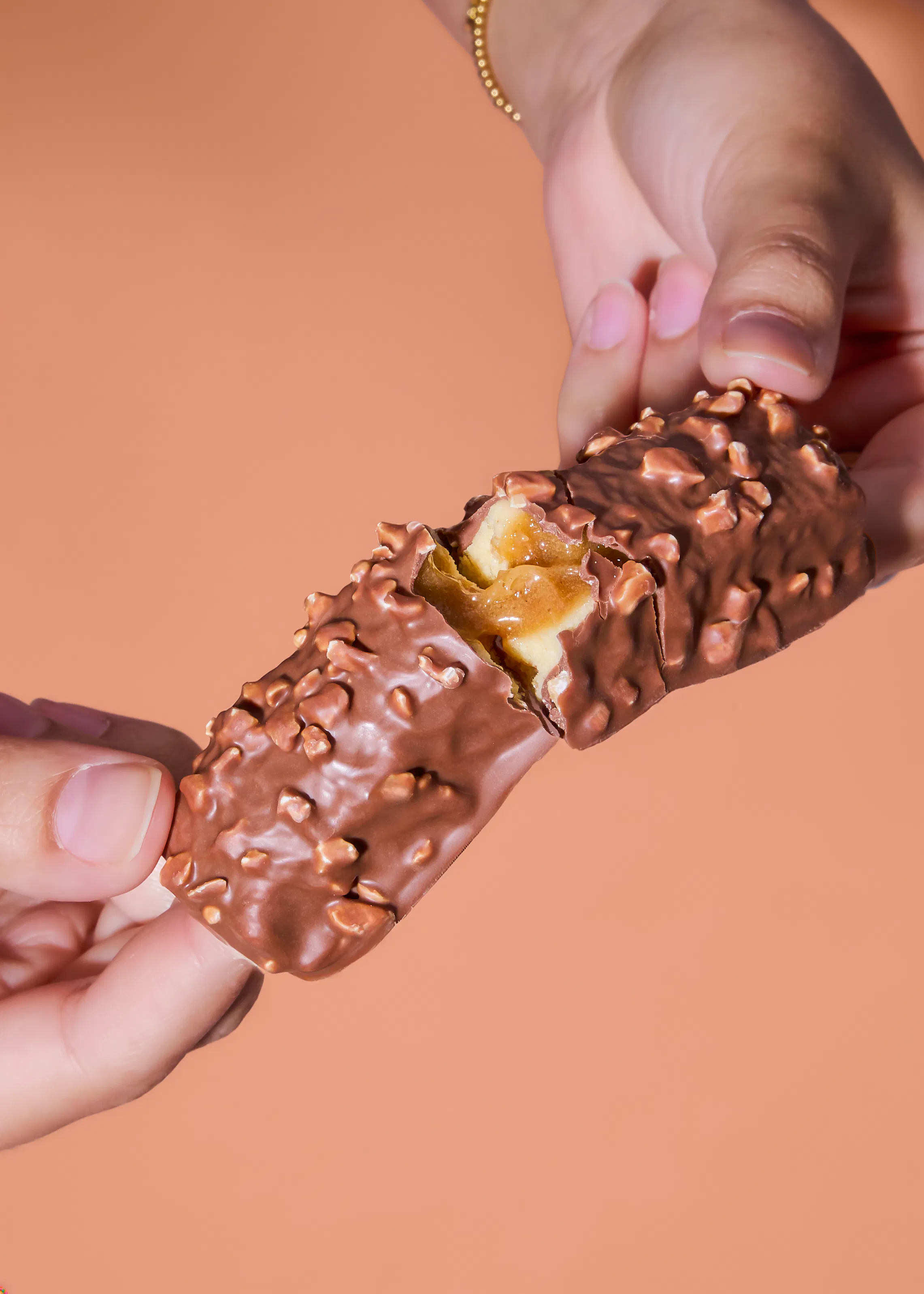 due mani tengono una barretta di cioccolato ricoperta di noci, spezzata a metà per rivelare un ripieno cremoso e caramellato.