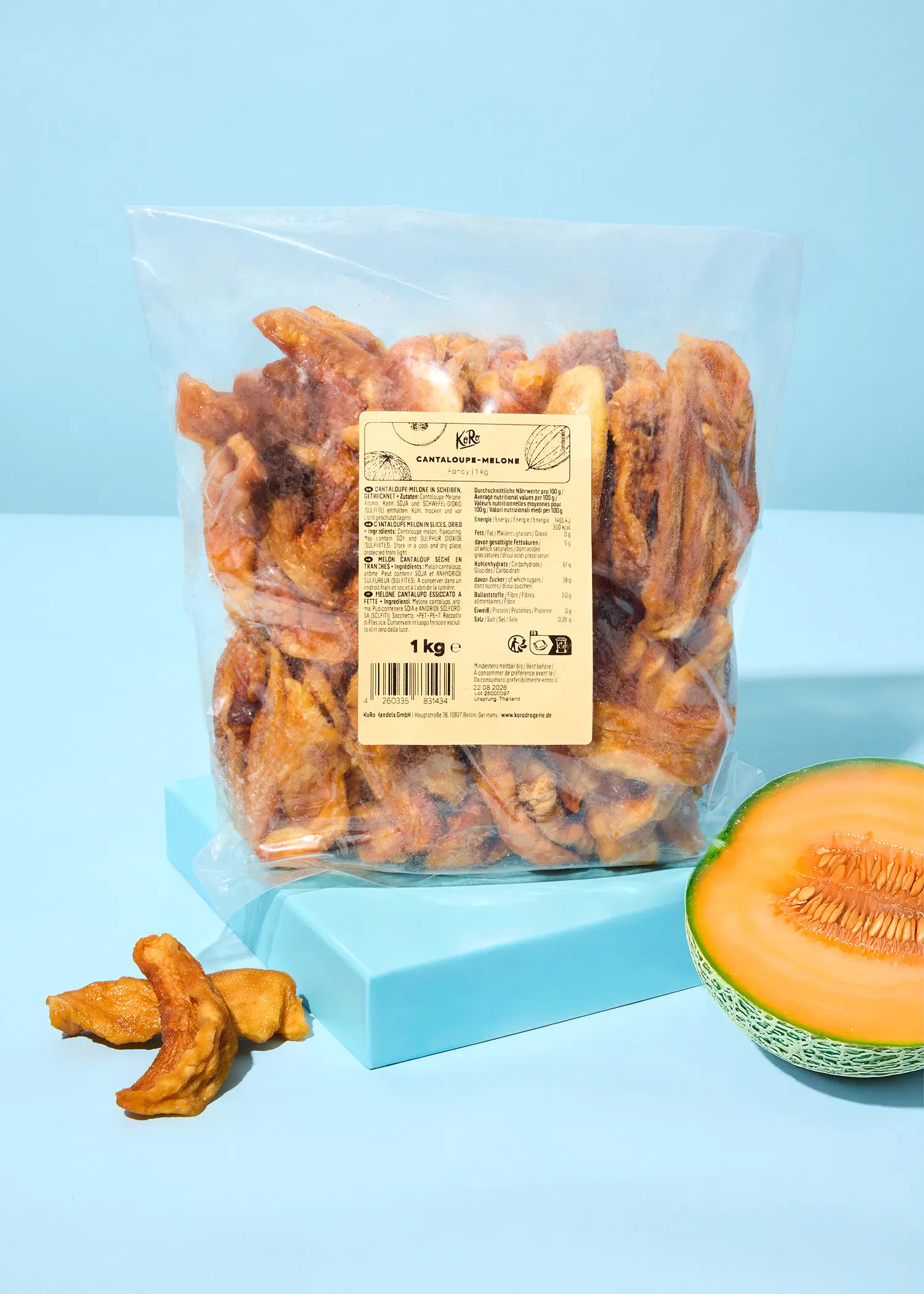 a clear plastic bag of dried cantaloupe melon slices sits on a light blue surface next to a fresh half cantaloupe and a few loose dried pieces.