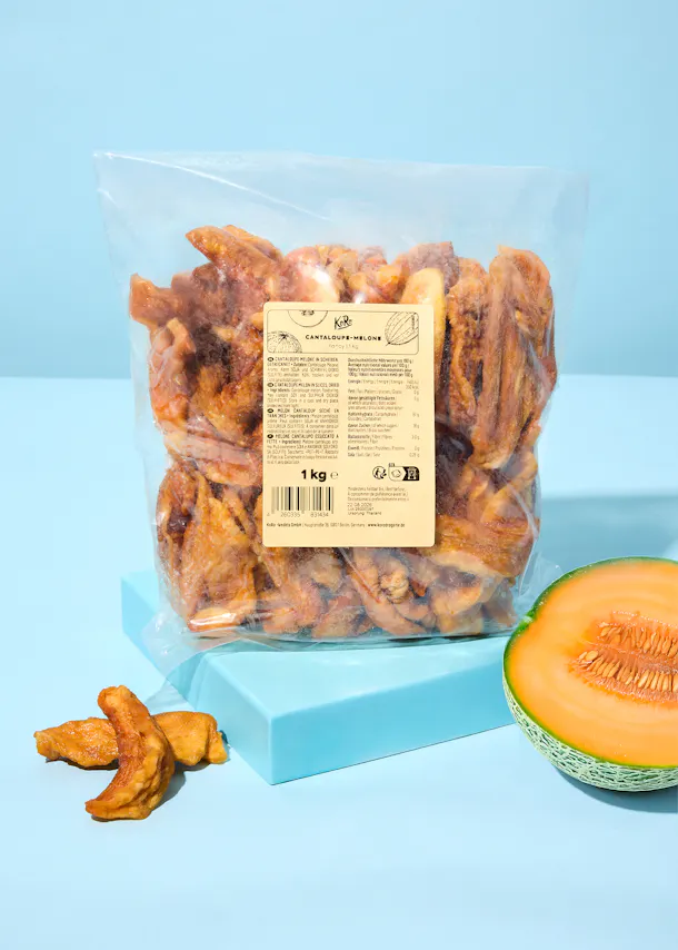 a clear plastic bag of dried cantaloupe melon slices sits on a light blue surface next to a fresh half cantaloupe and a few loose dried pieces.