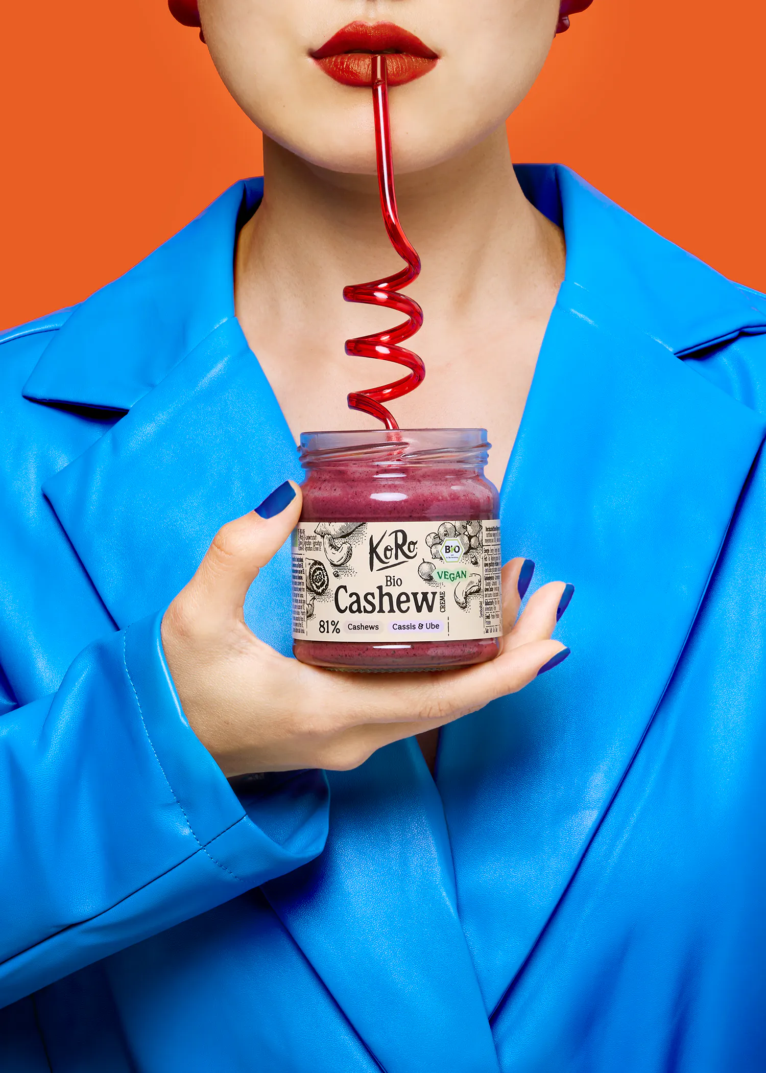 eine person mit rotem lippenstift und blauer jacke trinkt mit einem roten spiralförmigen strohhalm aus einem glas koro bio cashew creme cassis & ube.