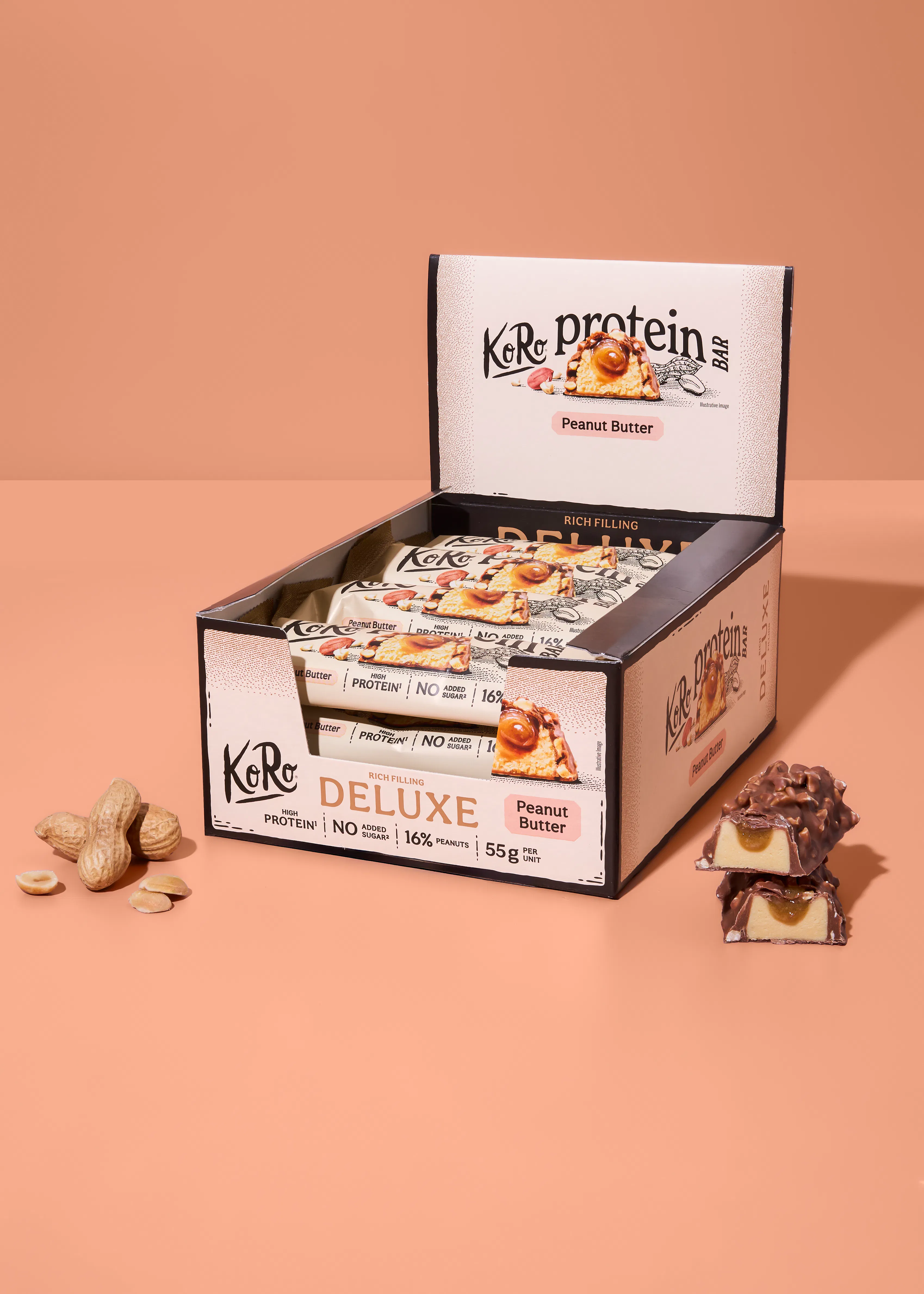 una caja expositora abierta de barritas de proteína koro deluxe sabor cacahuete, con varias barritas dentro y una barrita cortada por la mitad mostrando su relleno cremoso, junto a cacahuetes con cáscara y pelados.