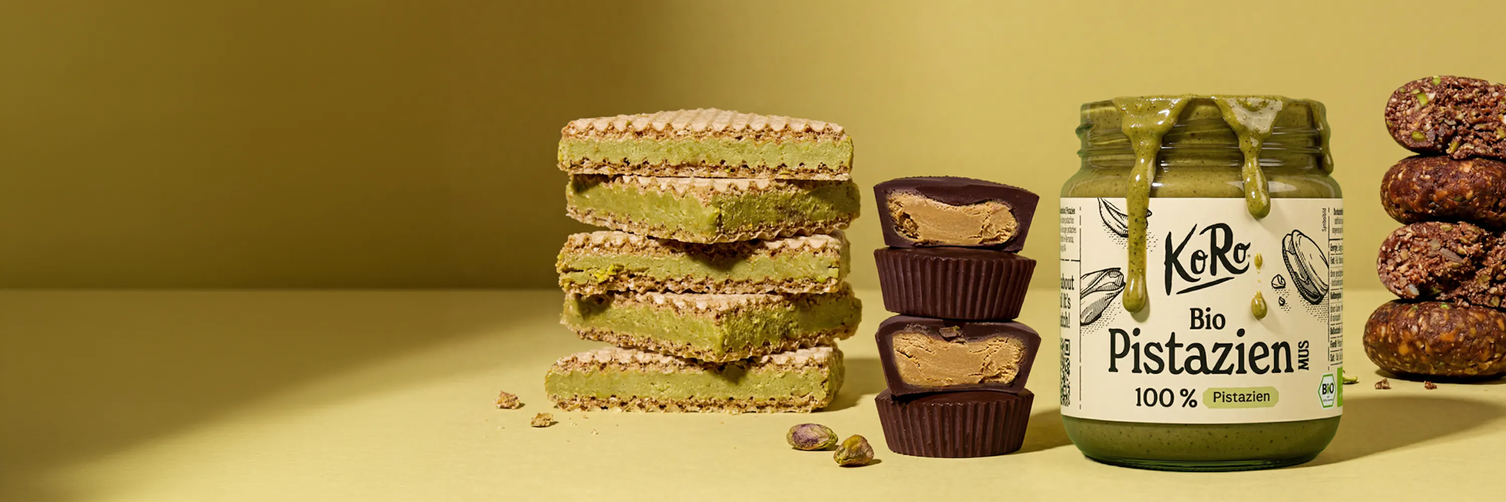 ein stillleben mit einem stapel pistazienwaffeln, schokoladenbechern mit erdnussbutterfüllung, einem glas pistazienmus und energiekugeln auf einem gelbgrünen hintergrund.