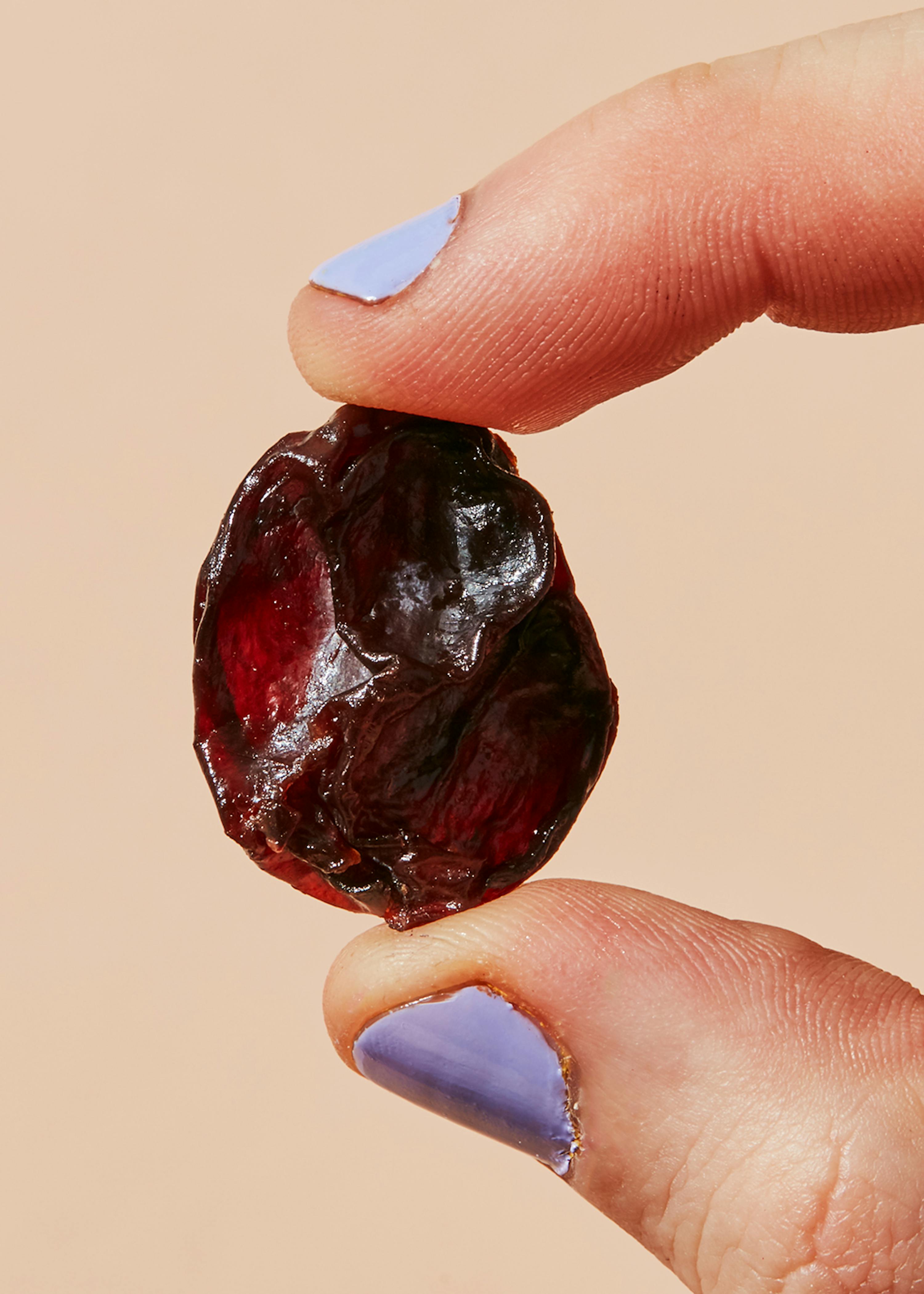 eine Person mit lila Nägeln hält eine Kirsche zwischen ihren Fingern