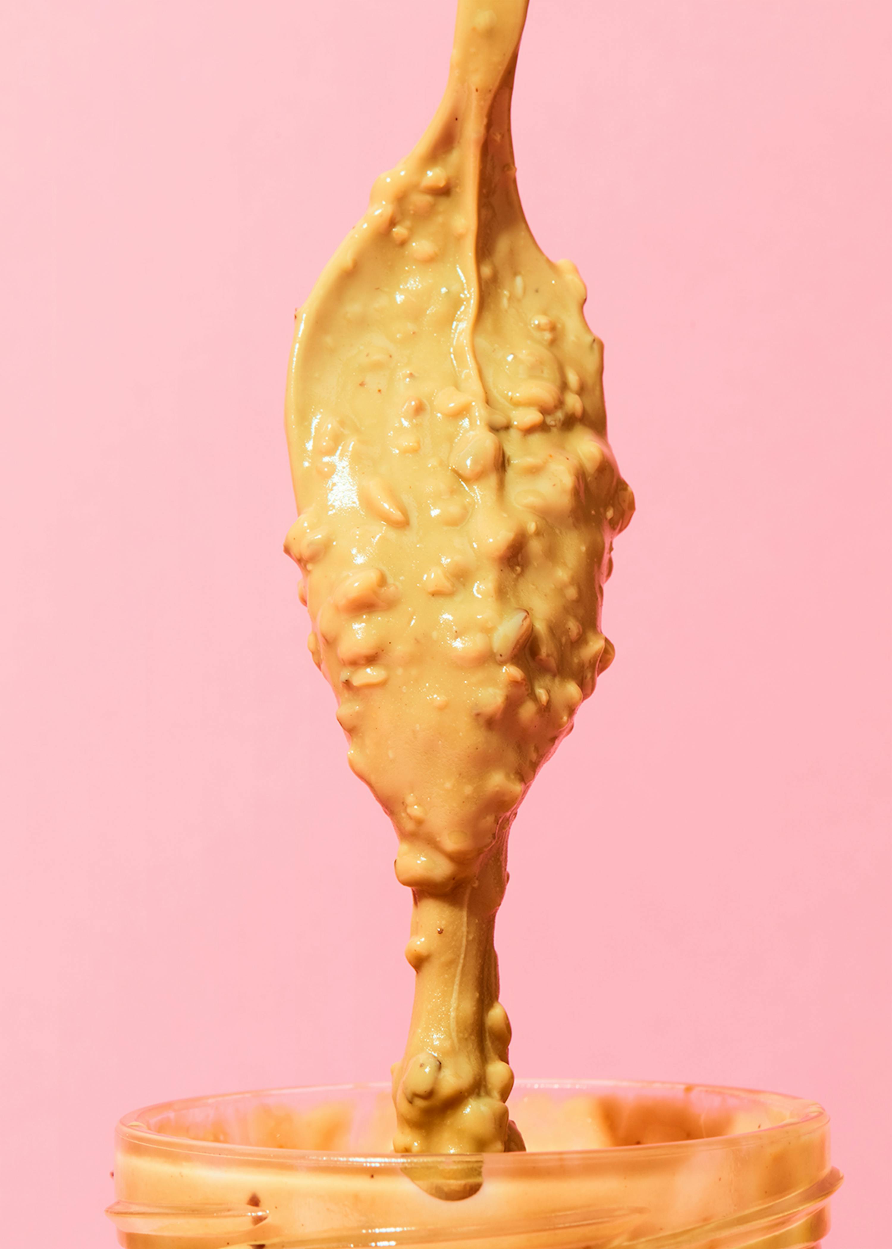 un cucchiaio di burro di arachidi viene versato in un barattolo su sfondo rosa