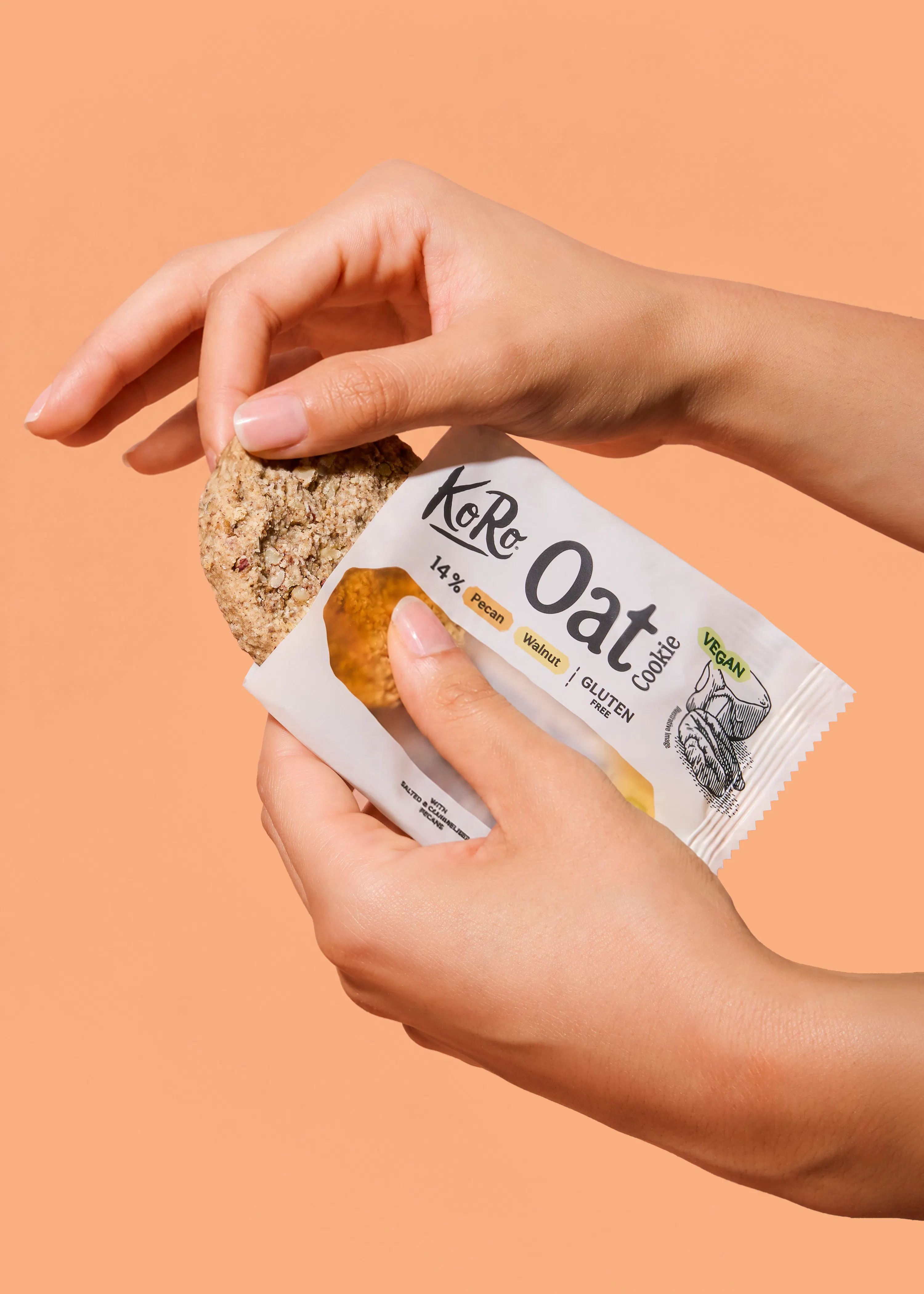 hände halten einen koro oat cookie mit pekannüssen vor einem beigen hintergrund.