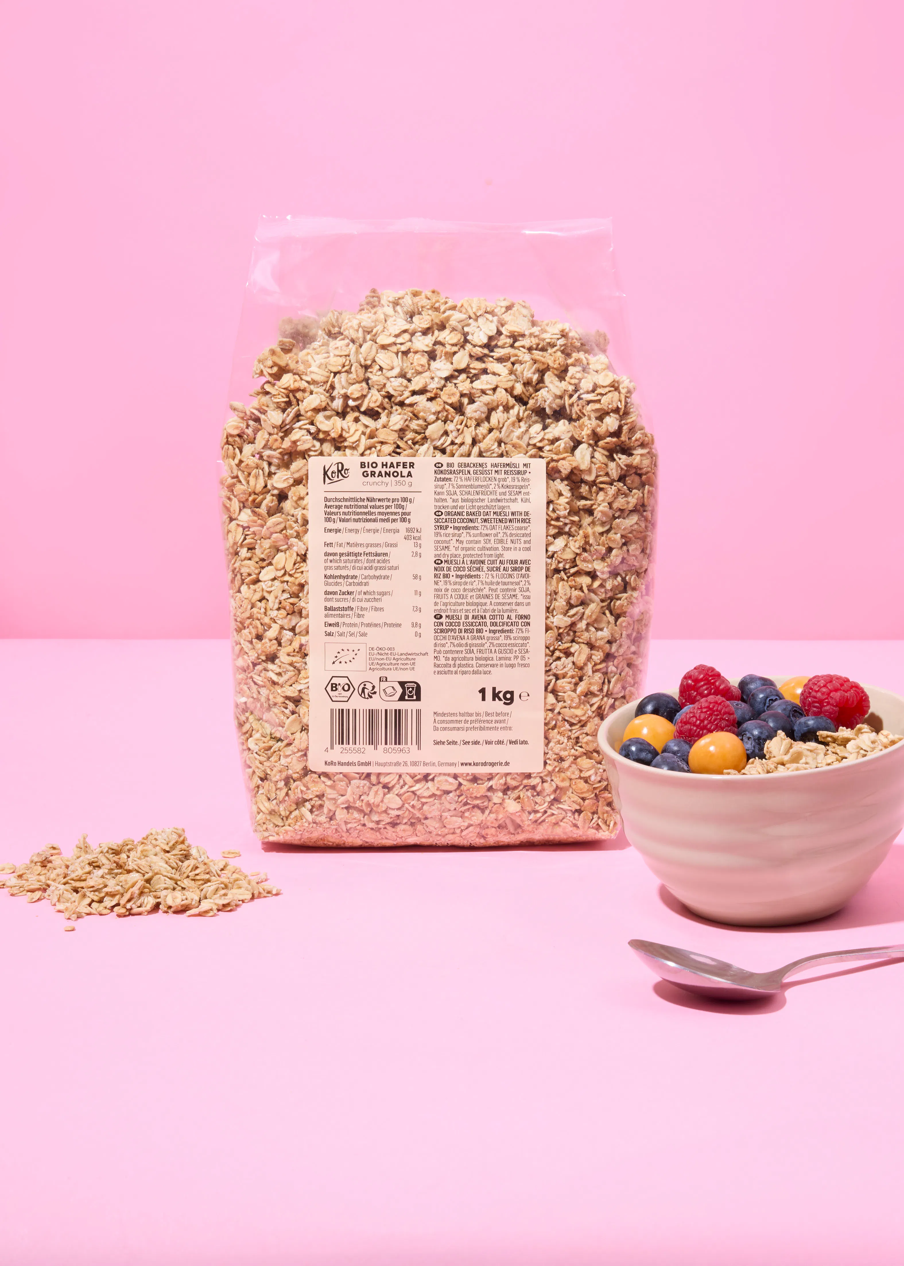 un grand sac de granola koro de 1 kg, une petite pile de flocons d'avoine et un bol de granola avec des baies et des physalis, accompagnés d'une cuillère, sur un fond rose.
