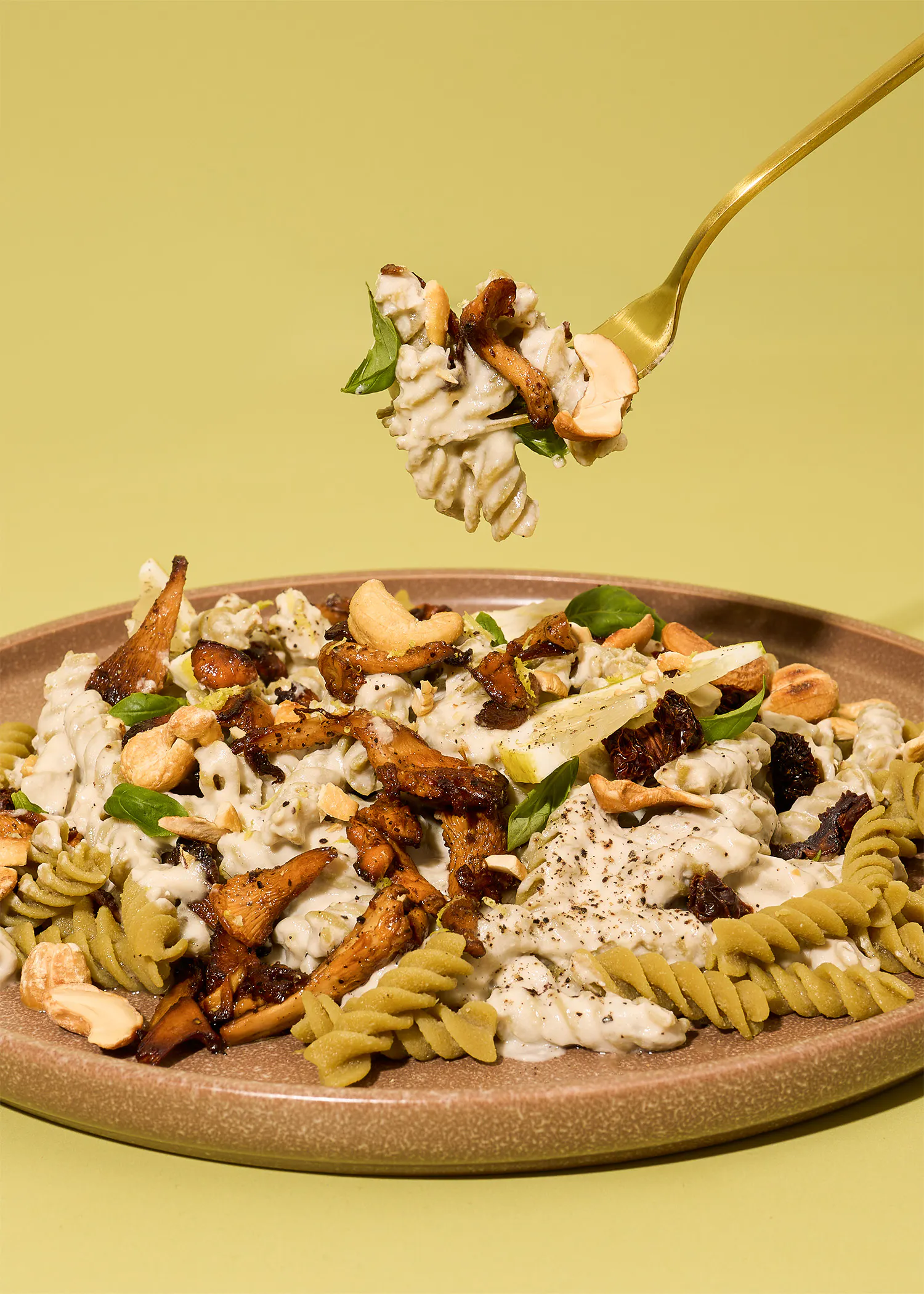 a gold fork lifts a bite of creamy fusilli pasta topped with roasted mushrooms, cashews, and fresh basil leaves from a plate against a light green background.
