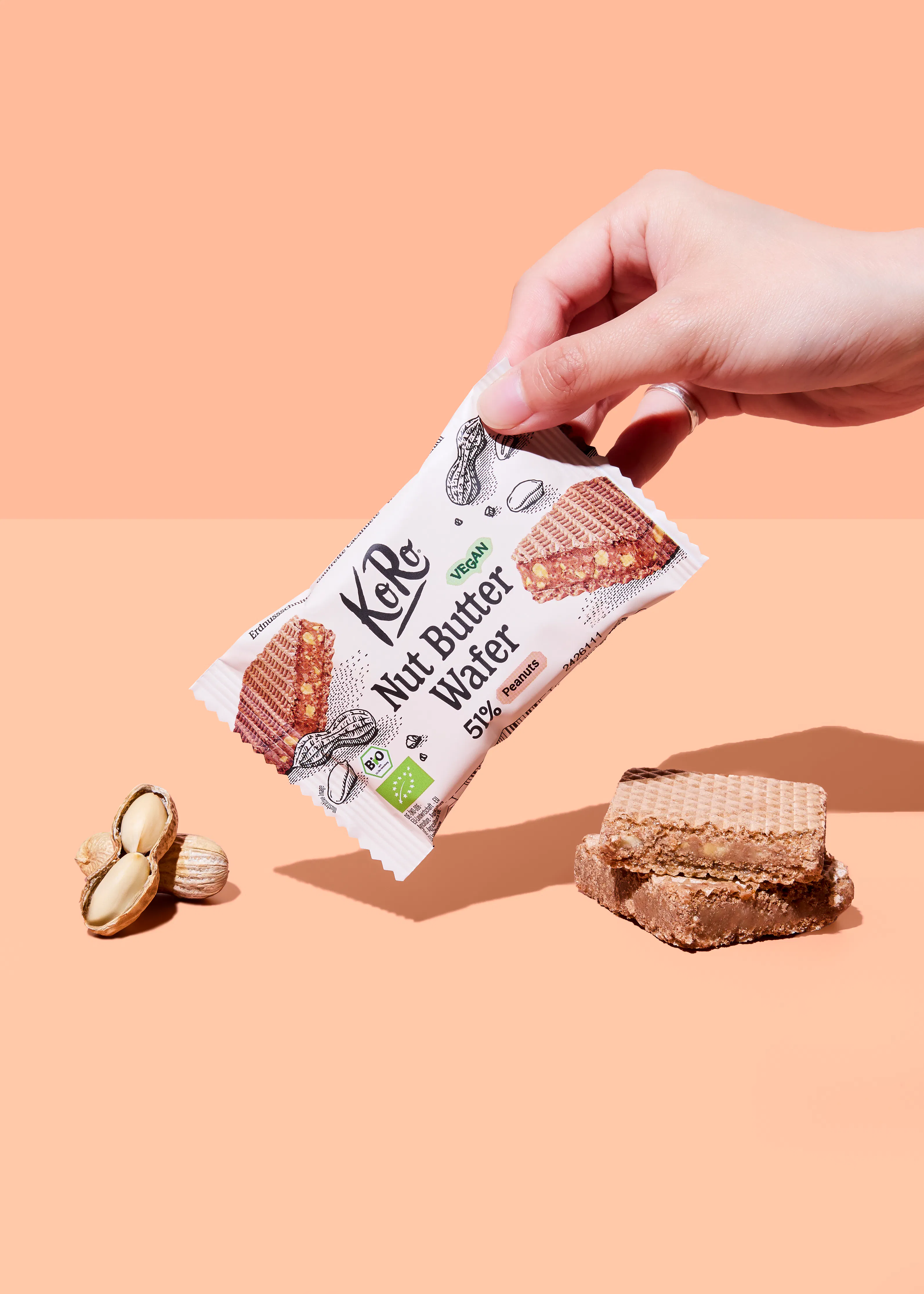 eine hand hält eine weiße packung eines koro nussbutter-waffelriegels, daneben liegen erdnüsse und zwei waffelstücke auf einem pfirsichfarbenen hintergrund.