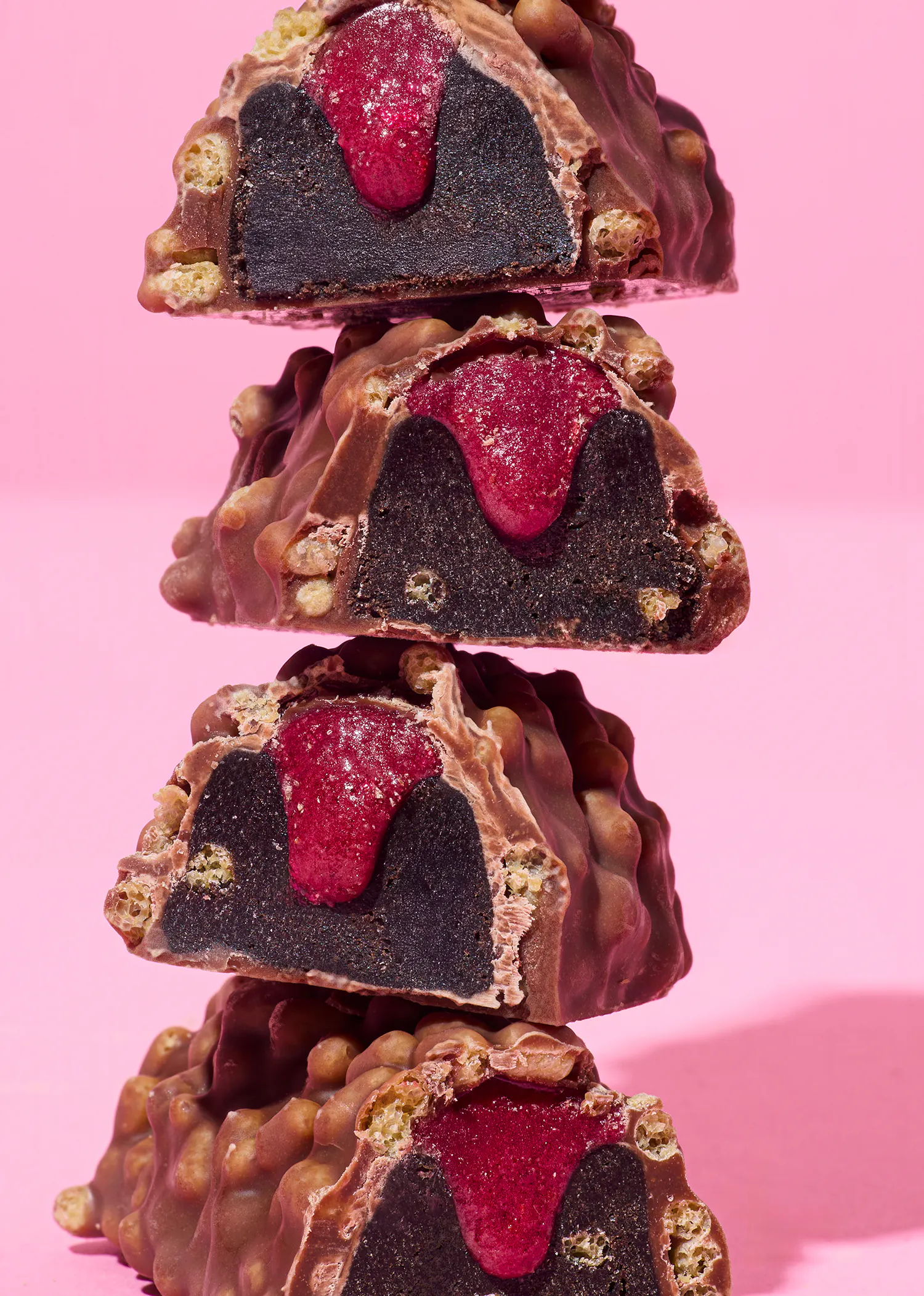 four pieces of chocolate with a raspberry filling are stacked on top of each other on a pink background