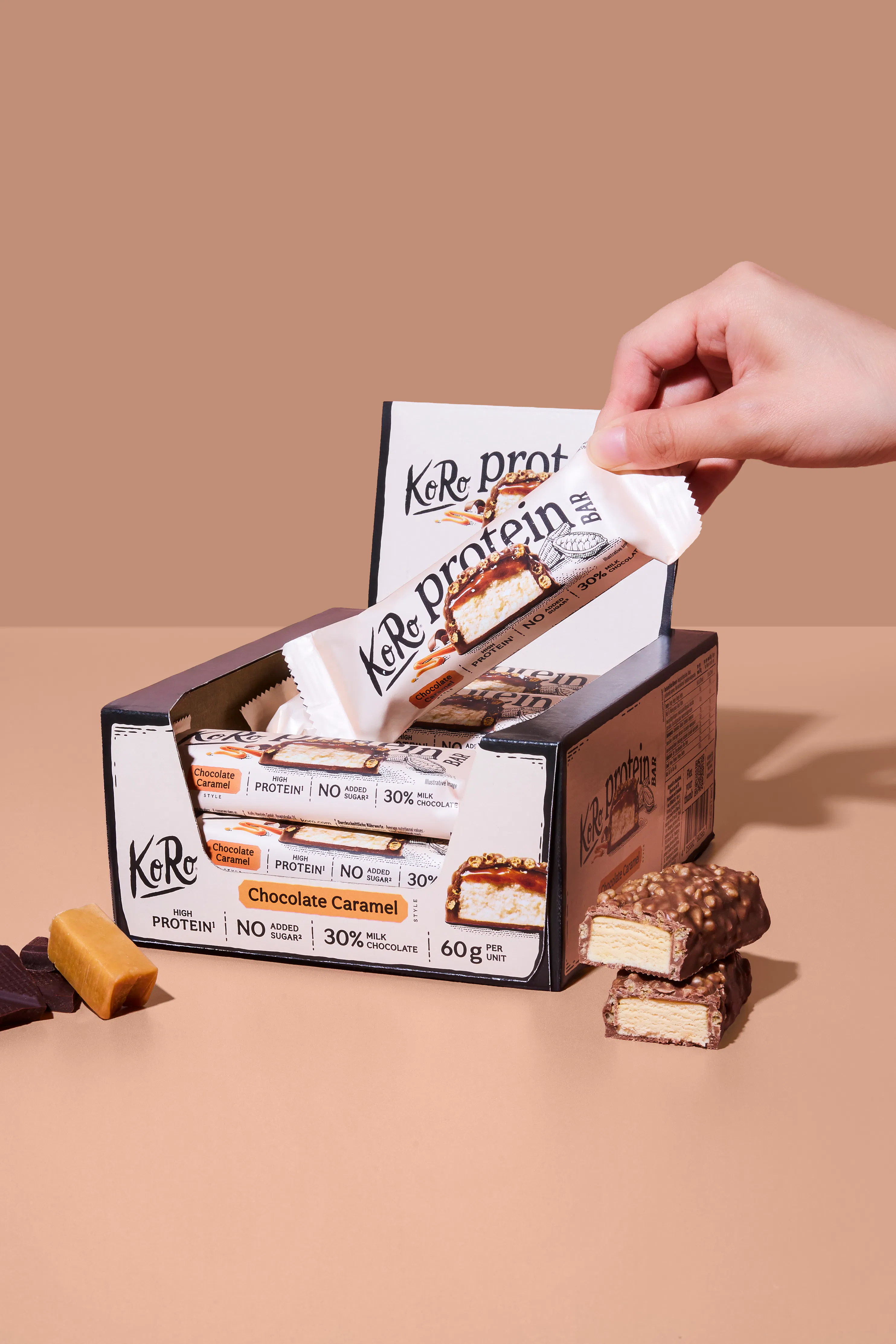 une main prend une barre protéinée koro saveur chocolat caramel d'une boîte, tandis que d'autres barres coupées et un morceau de caramel sont visibles sur un fond beige.
