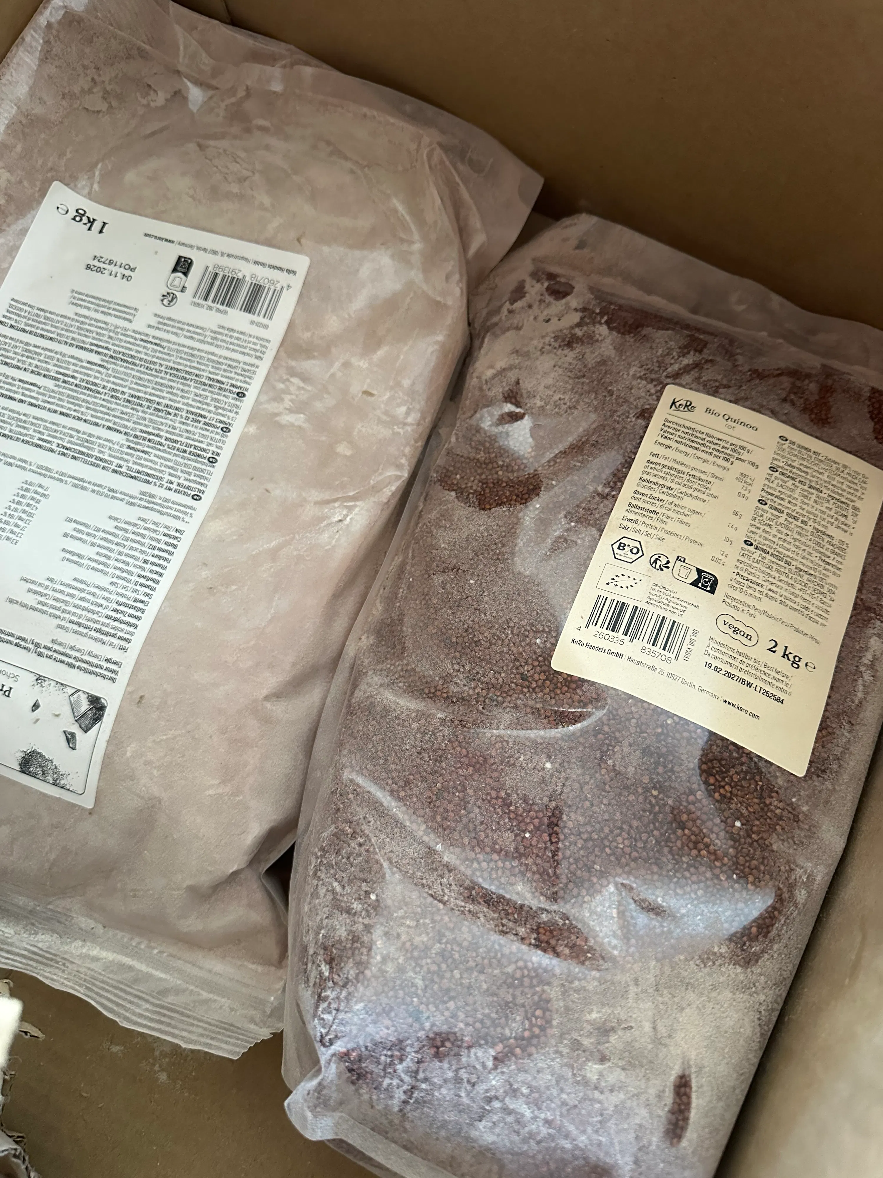 zwei große plastikbeutel mit lebensmitteln, darunter roter quinoa und proteinpulver, liegen in einem karton.
