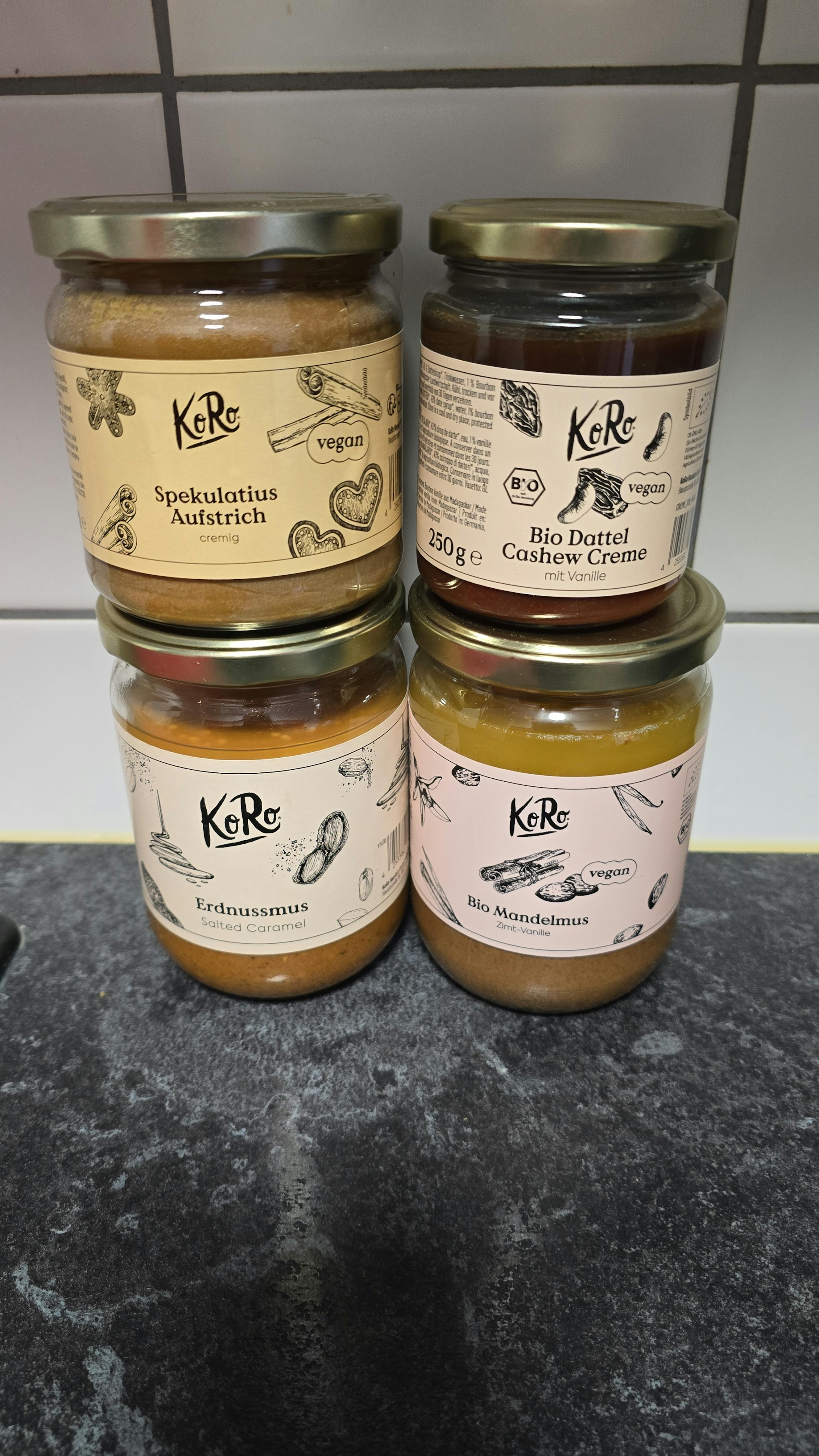 quatre pots de pâtes à tartiner véganes de la marque koro, incluant crème de dattes et noix de cajou, beurre d'amande cannelle-vanille, pâte à tartiner spéculoos et beurre de cacahuète caramel salé, posés sur un comptoir sombre.