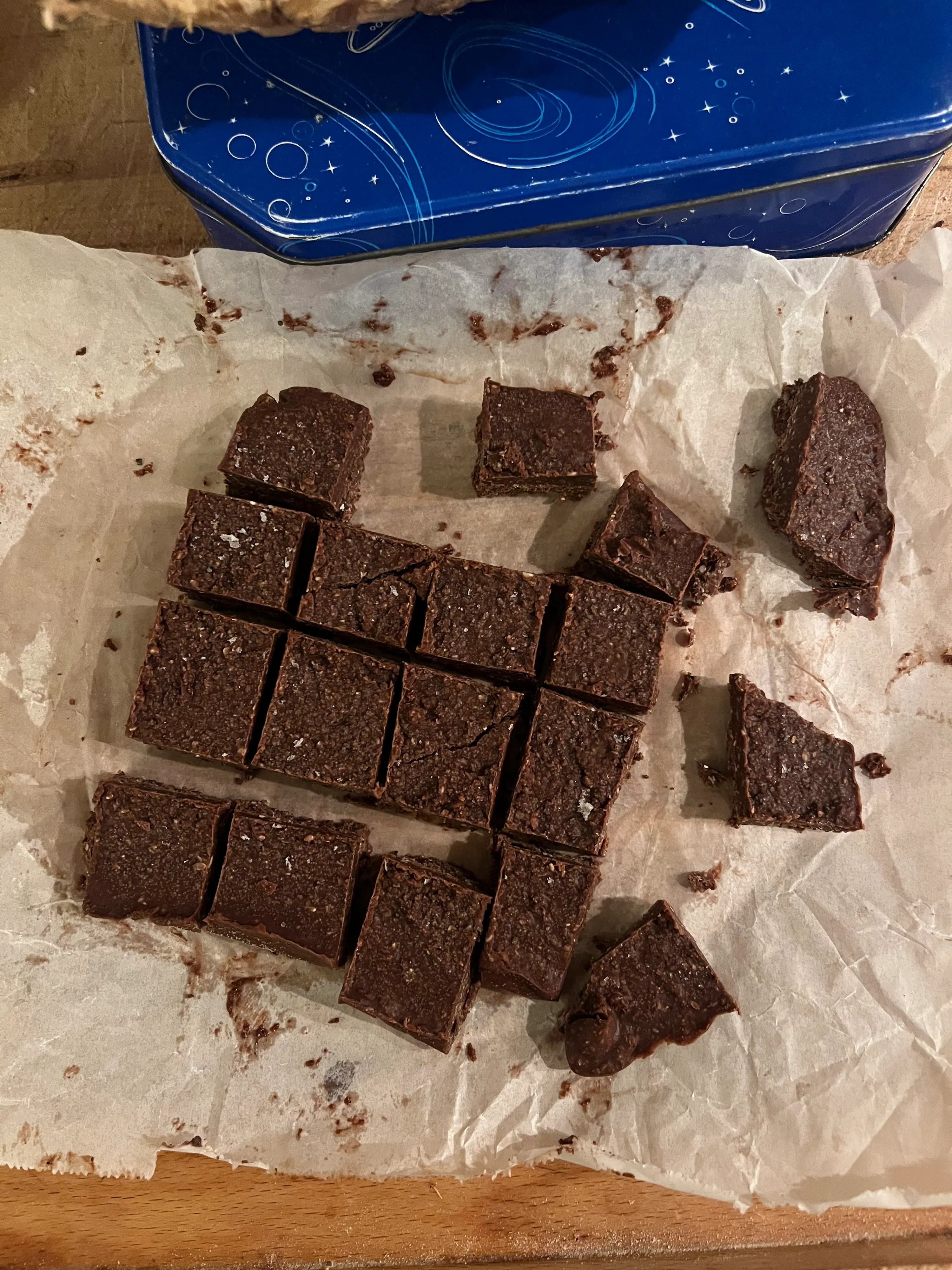 fudge al cioccolato scuro tagliato a pezzi su carta da forno accartocciata, con una scatola blu a sinistra.